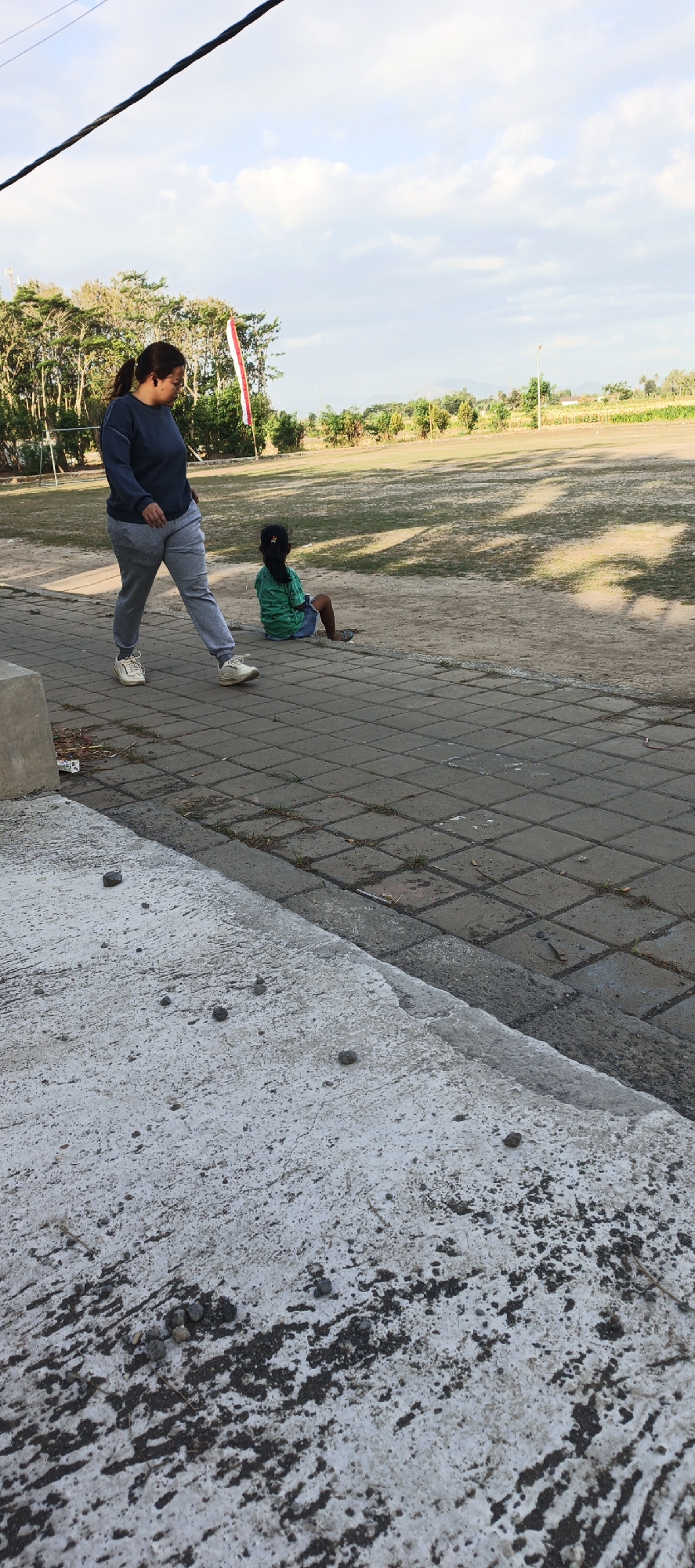 In Kasiyan Timur, Indonesia, a woman in a dark blue long-sleeved shirt and grey sweatpants walks on a paved walkway. She is facing left and looking down. A child, wearing a green and black striped shirt and blue shorts, sits on the dirt ground to the right of the walkway, facing away from the viewer. Behind them, a grassy field is visible with trees and a red and white flag. The sky is partly cloudy.