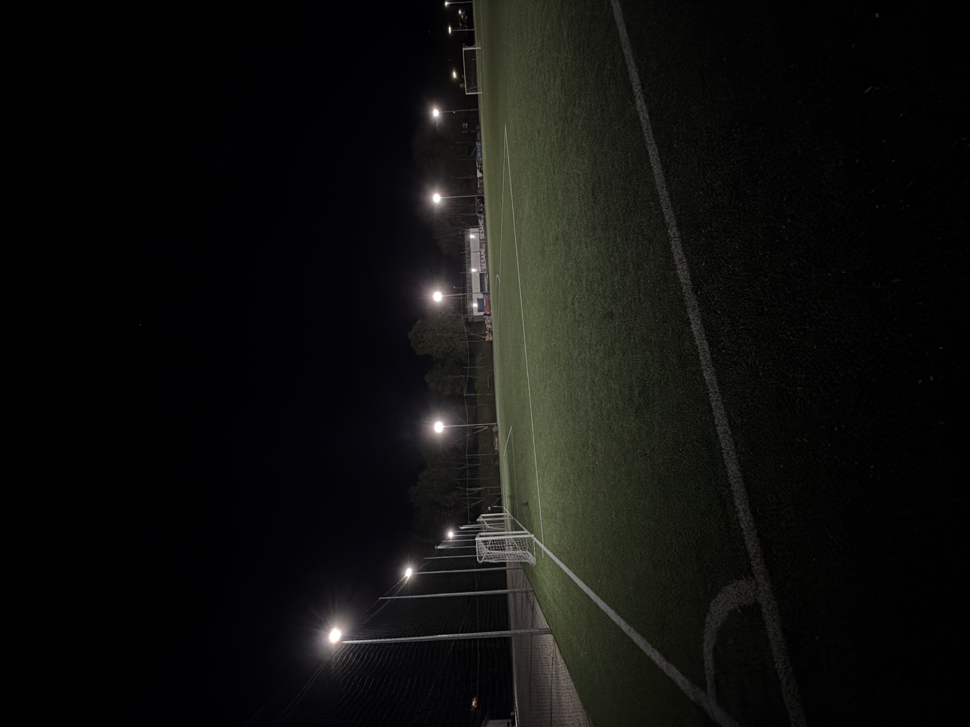 The image displays an empty football (soccer) field at night, brightly illuminated by a series of tall floodlights. The artificial green turf of the pitch is clearly visible, marked with crisp white lines indicating the sideline, a goal area line, and a corner arc. A white football goal, complete with netting, stands towards the end of the field.
The powerful lights on the poles cast a stark glow on the playing surface, creating a strong contrast with the dark, unlit sky that occupies a significant portion of the left side of the frame. A fence, possibly a chain-link, runs along the length of the field, behind which are dark silhouettes of trees. In the mid-ground, behind the lights and fence, several advertising boards or banners are visible, clearly displaying the word "SESAME" repeated horizontally.
There are no people present on the field or in the immediate vicinity, suggesting the scene is captured either before or after a game or practice session, or when the field is simply dormant. The overall atmosphere is one of stillness and anticipation. The time of day is night, with a clear sky. The location is Pleven, Bulgaria.
