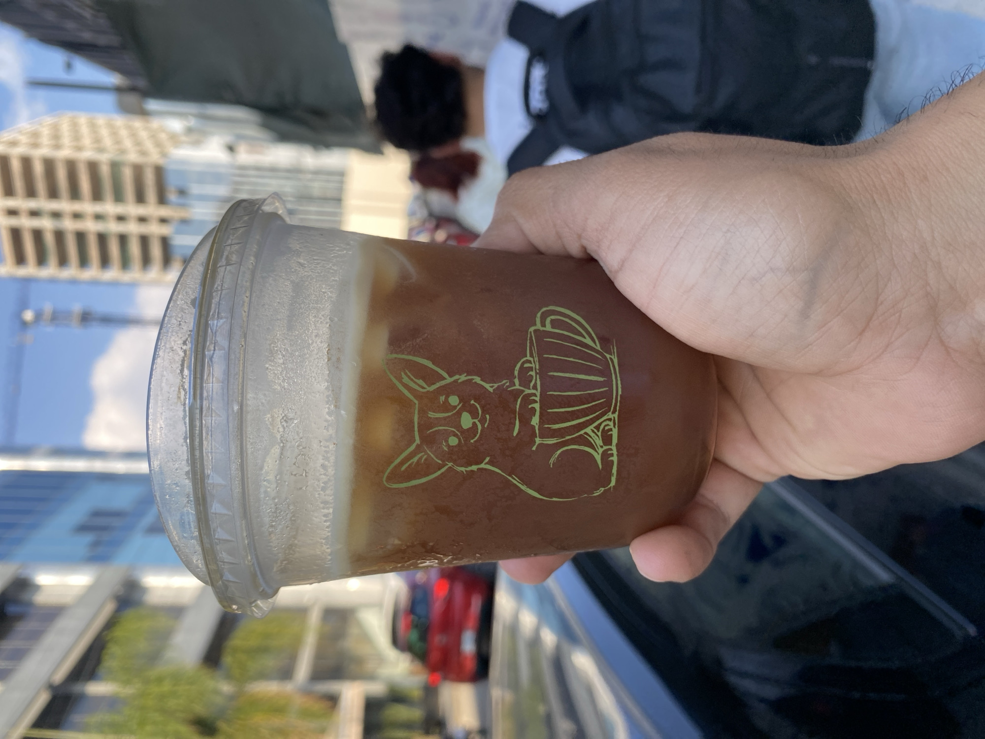 A hand holds a clear plastic cup filled with a brown beverage and a layer of white foam on top. The cup has a green cartoon drawing of a dog holding a mug on its side. The cup appears to be cold as condensation is visible on the outside. Buildings are visible in the background, suggesting an urban setting. A red car is visible in the bottom of the cup's reflection. Part of a black backpack and a person with dark hair are also visible in the background. The image does not provide enough information to determine the location is Puebla, Mexico.