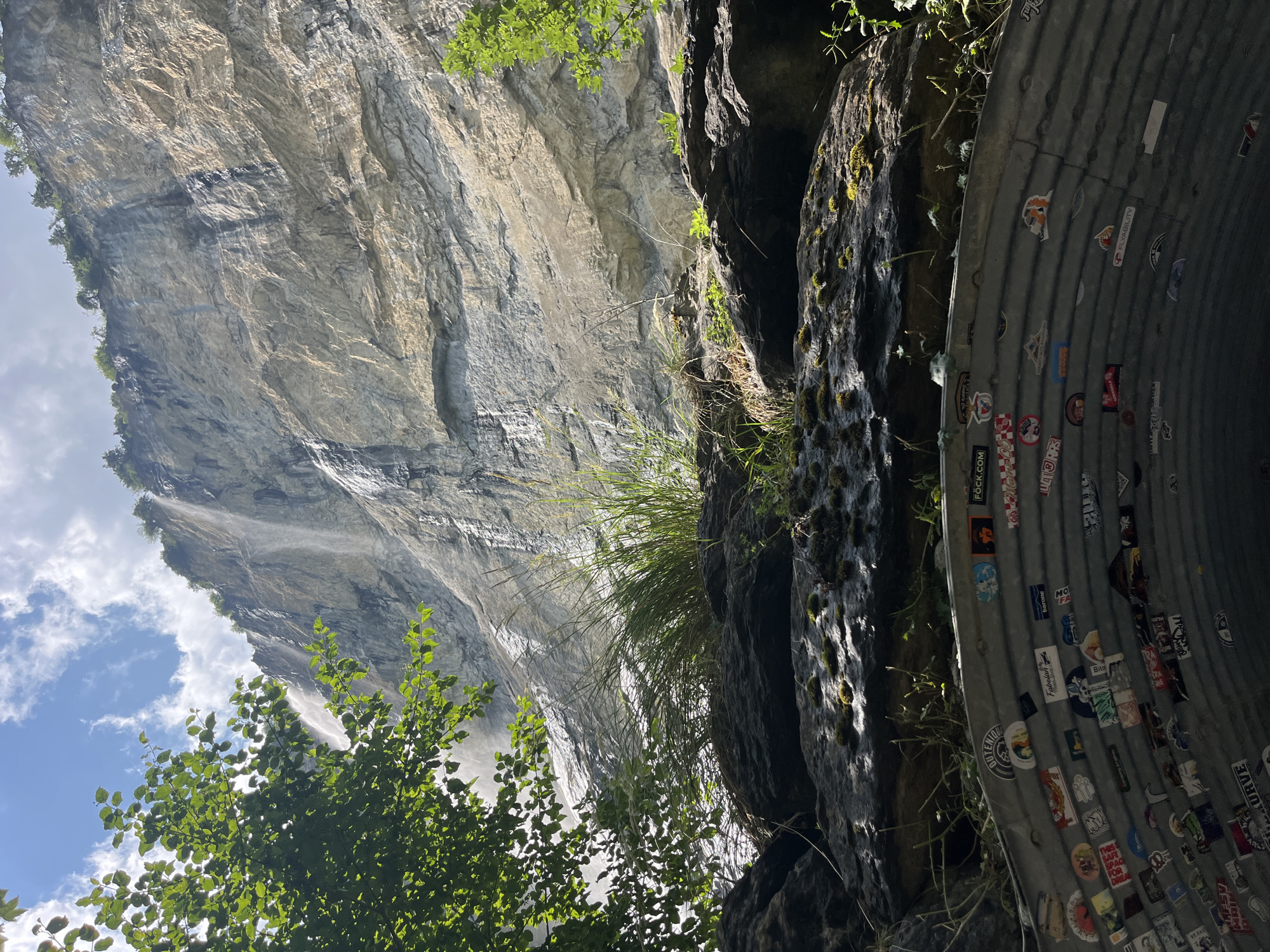 The image shows a view looking up towards a sheer, layered rock face with a thin waterfall cascading down its side. Lush green foliage frames the left side of the scene, and other green plants and moss grow from the rocky outcrops in the foreground. Below the rocky overhang, a curved, corrugated metal structure, likely part of a tunnel or drainage system, is visible. This structure is extensively covered in colorful stickers. The sky above is blue with scattered white clouds, and sunlight illuminates parts of the rock face. This view is characteristic of the area near the Staubbach Falls in Wengen, Switzerland, a popular tourist destination known for its dramatic waterfalls and alpine scenery. There are no people or animals visible in the image.