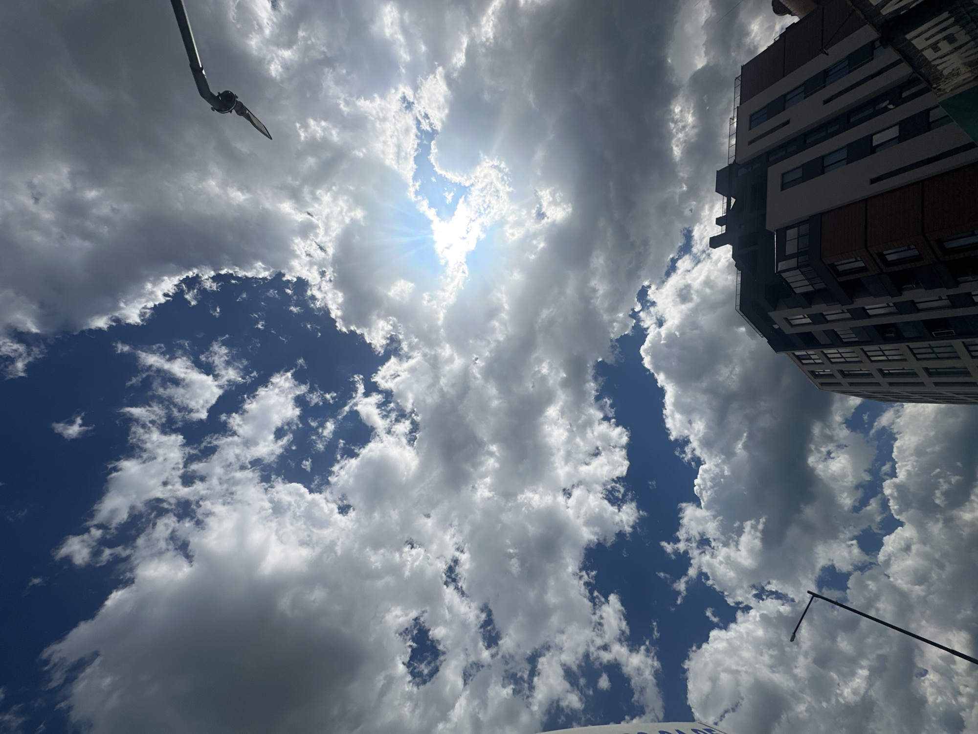 The image captures a dynamic sky over a modern apartment building in Sofia, Bulgaria. The sky is filled with a mix of white and dark gray clouds, with the sun breaking through in the upper right quadrant, casting rays of light. The building, predominantly beige and brick-colored, features numerous windows and balconies. In the foreground, the top of a lamppost is visible on the left, and a metallic arm extends from the top of the frame on the right. The perspective is looking upwards from a low angle.
