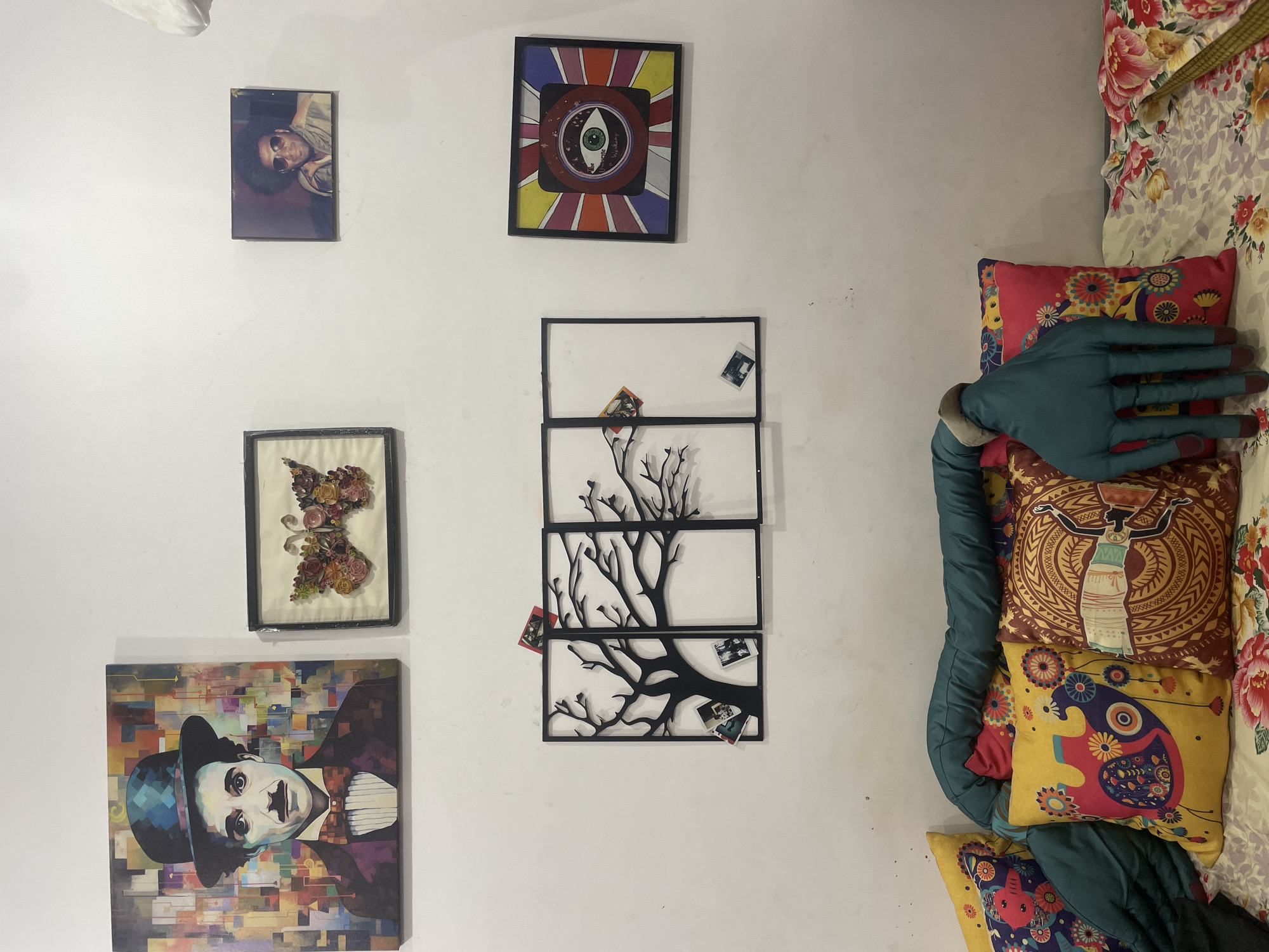 The image depicts a wall with various decorations. Five framed artworks are hanging on the wall: a portrait of a man with afro hair, an abstract image with an eye in the center, a flower design made of dried flowers, a tree design consisting of three stacked rectangular frames, and a portrait of Charlie Chaplin. To the right of the wall is a stack of pillows with colorful patterns and a green, padded body pillow with a large, blue hand extending from it. The scene appears to be indoors. There is no indication of the location being in Morjim, India.