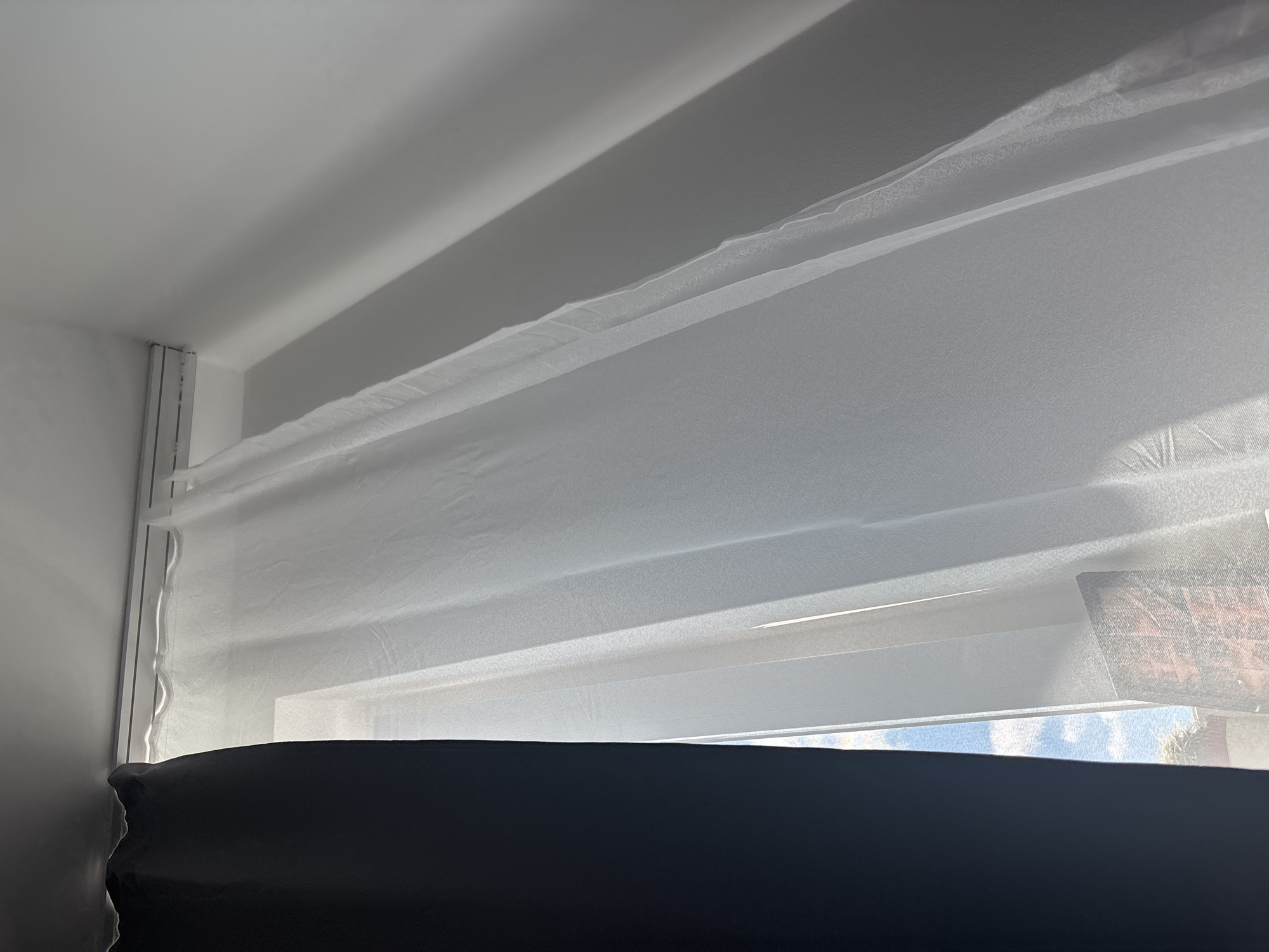 In Gebersdorf, Austria, an interior shot captures a window adorned with two layers of curtains. The left side is dominated by a thick, dark curtain, partially obscuring the view. To its right, a sheer white curtain billows, allowing sunlight to filter through and illuminate the room. The white curtain has a slightly textured surface, with subtle ripples and folds adding depth. In the lower portion of the frame, a glimpse of the sky and some greenery is visible through the sheer fabric, suggesting an outdoor scene beyond the window. The wall to the right of the curtains is painted a light color and shows a soft shadow cast by the window. The image is composed from a slightly angled perspective, looking upwards towards the top of the window frame and the ceiling.