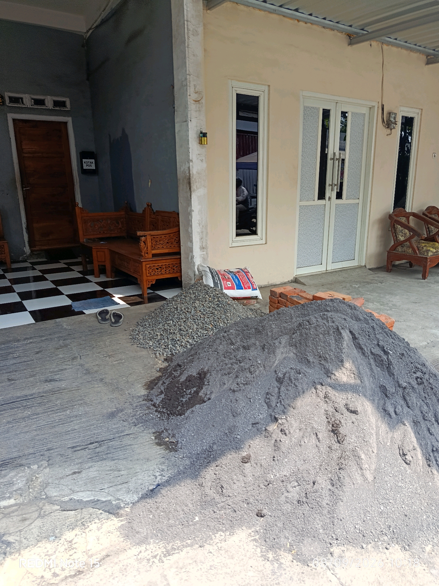 This is an exterior, eye-level, medium shot of a construction site in front of a home, likely located in Keboansikep, Indonesia. In the foreground, two large piles of construction materials are present: one of coarse gravel and another of fine, dark sand. To the left of the piles, a bag of cement is partially visible. A stack of red bricks lies between the piles and a beige-colored wall.
To the left of the construction materials, a tiled patio with a black and white checkerboard pattern is visible. On this patio, a wooden bench with intricate carvings is positioned. Further left, a dark brown wooden door is set into a dark gray wall. A black mailbox labeled "KOTAK POS" is mounted on this wall.
The house itself has a light beige exterior. Two tall windows with white frames and frosted glass flank a pair of white double doors with similar frosted glass panels. To the right of the house, a dark wooden chair with a patterned cushion is visible.
The ground is a gray concrete surface, showing some signs of wear and cracking. The roof of the structure appears to be made of corrugated metal, supported by visible metal beams. The lighting suggests it is daytime, with sunlight casting shadows. There are no people clearly visible in the scene.