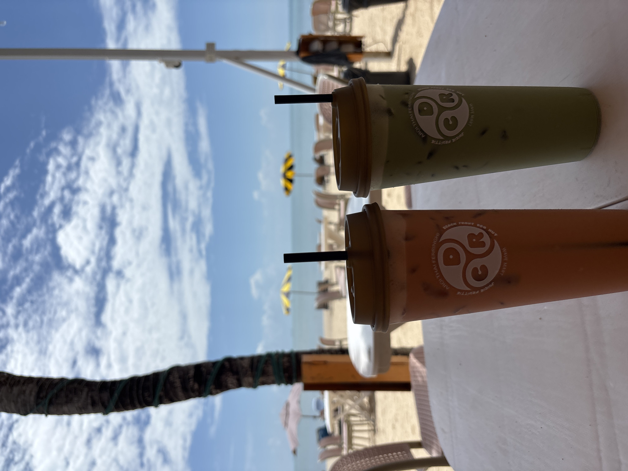 Two iced beverages, one orange and one green, sit on a white table at a beachside cafe. The orange drink is in the foreground on the left, and the green drink is to its right. Both cups have brown lids and black straws. A logo with the letters "DCR" inside circles is visible on both cups. In the background, beach chairs and umbrellas are set up on the sand, with the ocean and a cloudy blue sky visible beyond. A palm tree trunk, wrapped with green string lights, is on the left side of the image. The location appears to be a beach in George Town, Malaysia, based on the visible branding on the cups which reads "AROI THAI FER...". The overall scene depicts a relaxed, tropical setting.