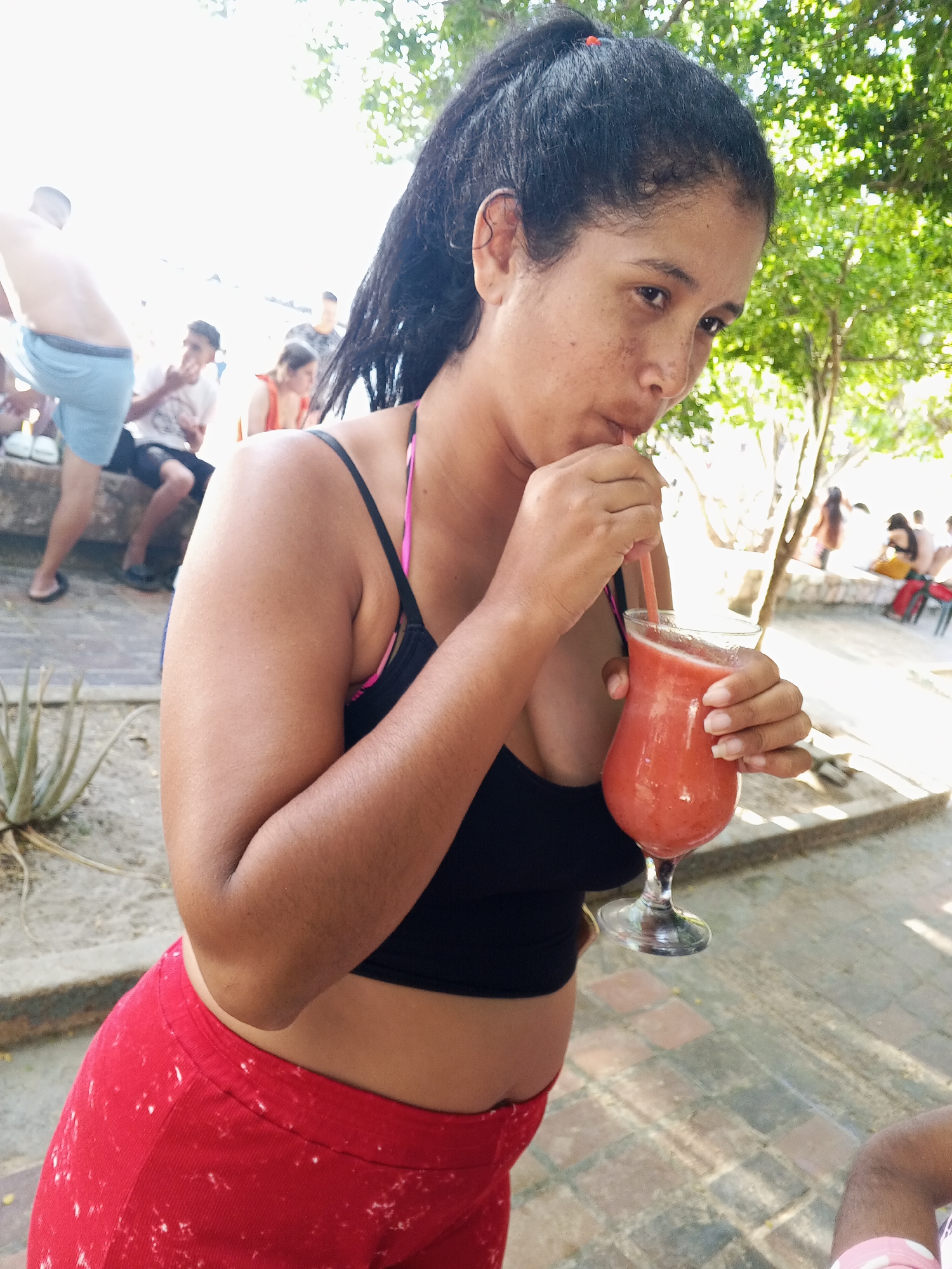 A woman with dark hair pulled back in a ponytail drinks a reddish smoothie with a straw from a stemmed glass. She is wearing a black crop top with pink straps and red pants with white speckles. The scene includes other people in the background, suggesting a public or semi-public location, possibly a recreational area near a beach in Pedro González, Venezuela, given the presence of trees and a view of people in swim attire. The ground appears to be paved with stone or brick.