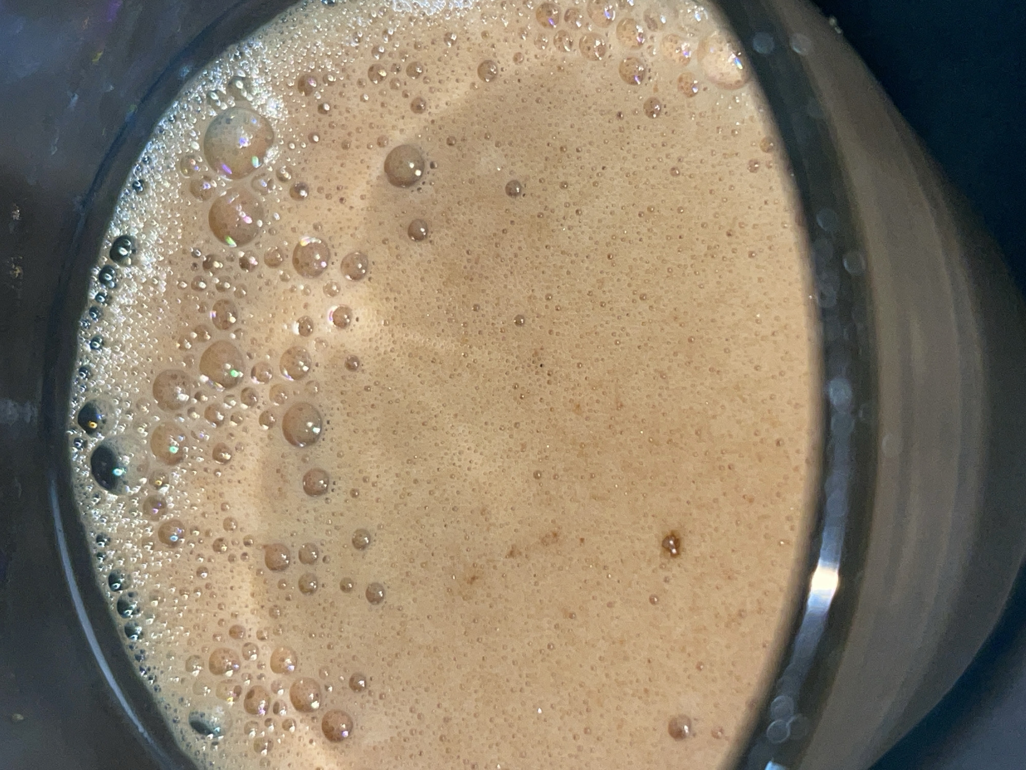This is a close-up, overhead shot of a dark-colored beverage with a thick layer of foam and bubbles. The beverage is a light brown color and appears to be in a dark, possibly glass, container. The foam has a creamy texture with many small bubbles, and some larger, iridescent bubbles are visible, catching the light. The lighting creates highlights on the surface of the foam and on the rim of the container. The image is focused on the beverage itself, with the background being out of focus and dark. There is no information in the image to suggest a location in Garut, Indonesia.