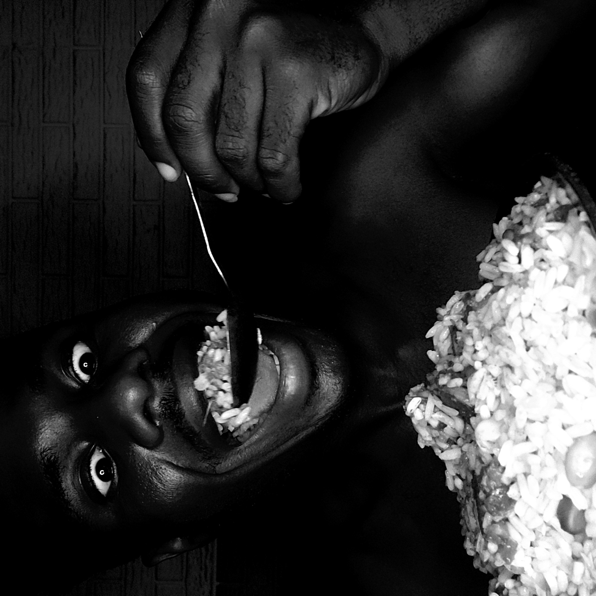 A stark, black and white photograph depicts a man eating a forkful of rice. He is on his side, with his face close to the camera. His eyes are open wide, and his mouth is open with rice visible on the tongue. In his right hand, he holds a fork with rice being guided toward his mouth. A plate or bowl containing more rice is beside his chest. The background is dark, suggesting a dimly lit room, and includes a hint of a textured wall. The image is turned ninety degrees counter-clockwise. Narayi, Nigeria, is not discernible from the image.