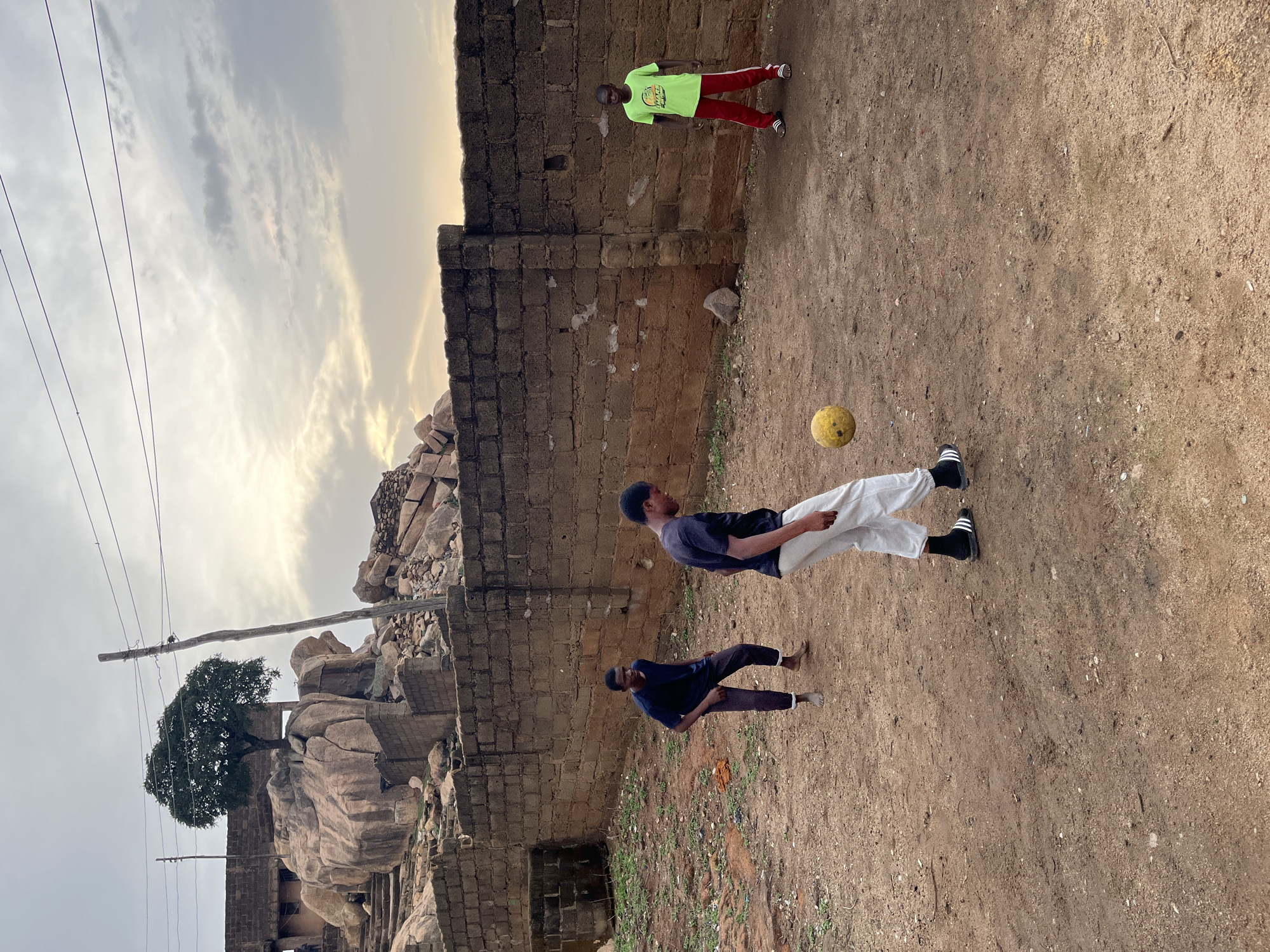 The image shows an outdoor scene in Jos, Nigeria. The environment features a dirt ground, a rough brick wall, and a rocky outcrop with a tree on top, utility poles and power lines are strung across the sky. Three people are present. One person is in the center of the image with their legs bent, in the background, one is standing against the wall. The third is on the ground with bent knees and arms. A yellow soccer ball is on the ground near the first person.