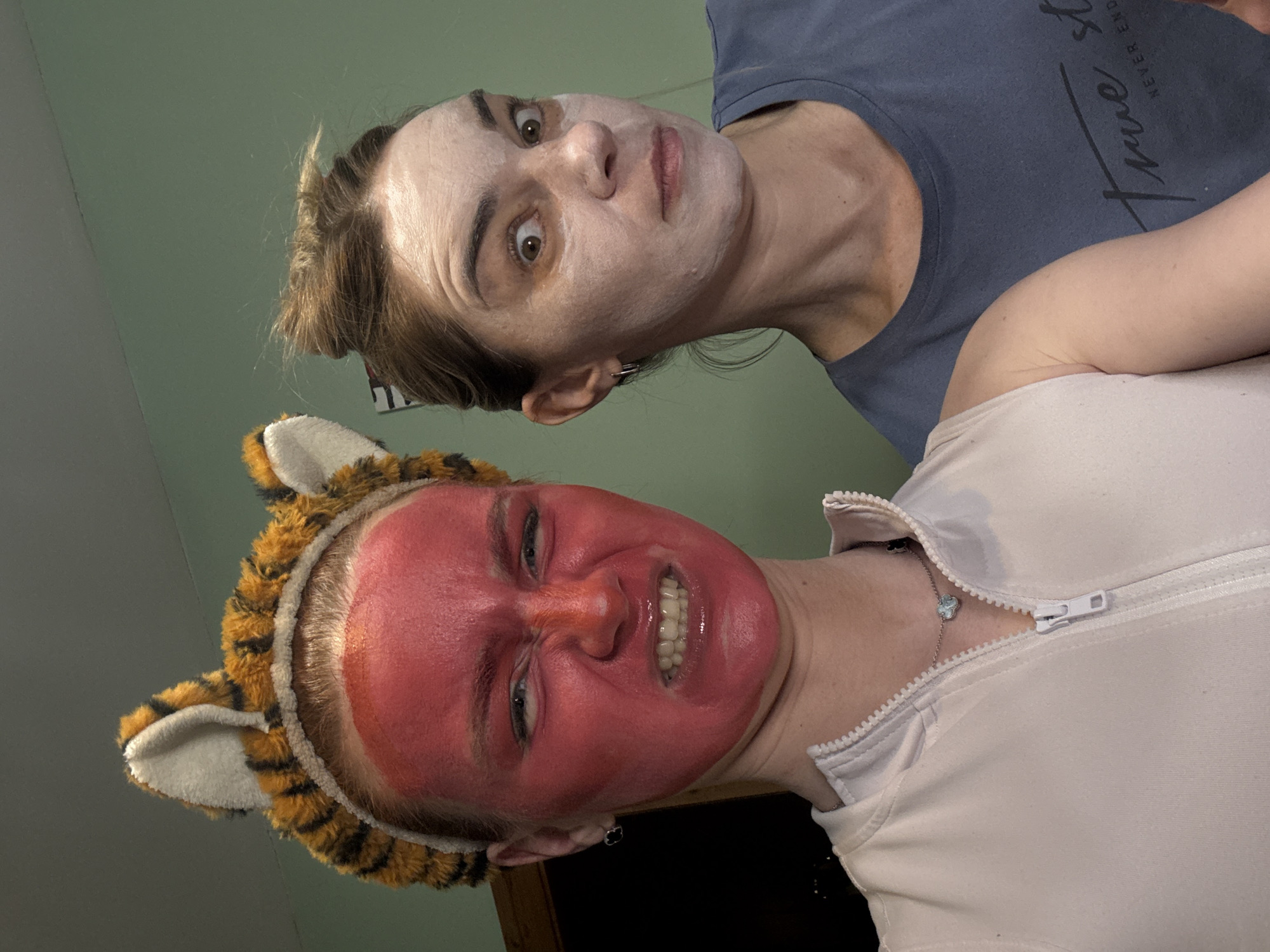 Two women are posing for a picture. The woman in the foreground is wearing a tiger-ear headband and has a bright red face mask on. She is grimacing and showing her teeth. She is wearing a light-colored zipped top and a silver necklace. The woman in the background has a white face mask on and is wearing a dark blue t-shirt with words on it. The background is a pale green wall. The location in the city of Sinyavino, Russia is unknown.