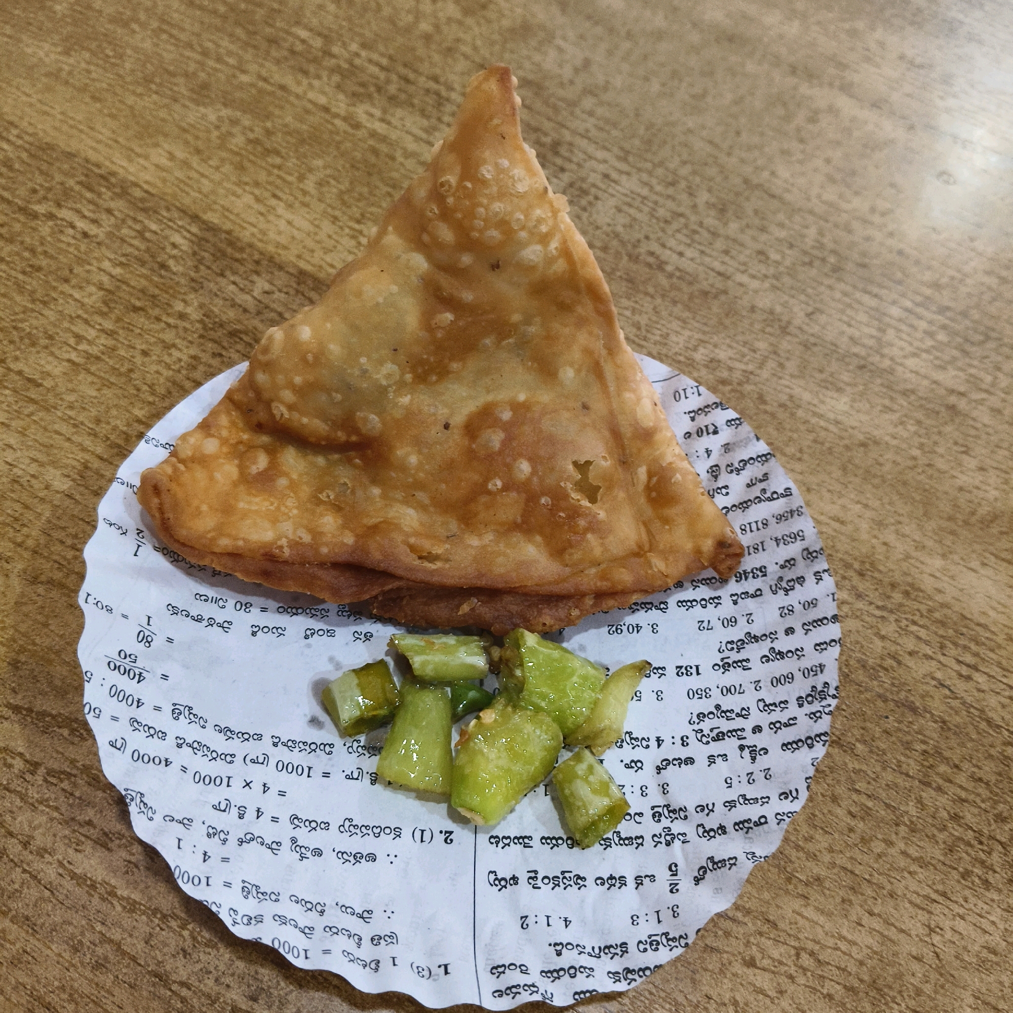 A close-up, overhead shot shows a single, golden-brown samosa placed on a piece of white paper with text in Telugu. The samosa, a triangular fried pastry, has a slightly uneven surface with visible air bubbles and some darker brown spots indicating it is cooked. To the right of the samosa, on the same paper, are several pieces of chopped green chili peppers, glistening with oil. The paper appears to be a torn section of a newspaper or receipt, with lines of Telugu script and numerical figures visible. The background is a textured wooden surface, likely a table, with prominent wood grain patterns. The lighting is diffused, casting soft shadows. The image suggests a casual dining or snack setting, with the food presented as the primary subject. The city of Choppadandi, India, is mentioned as the location.