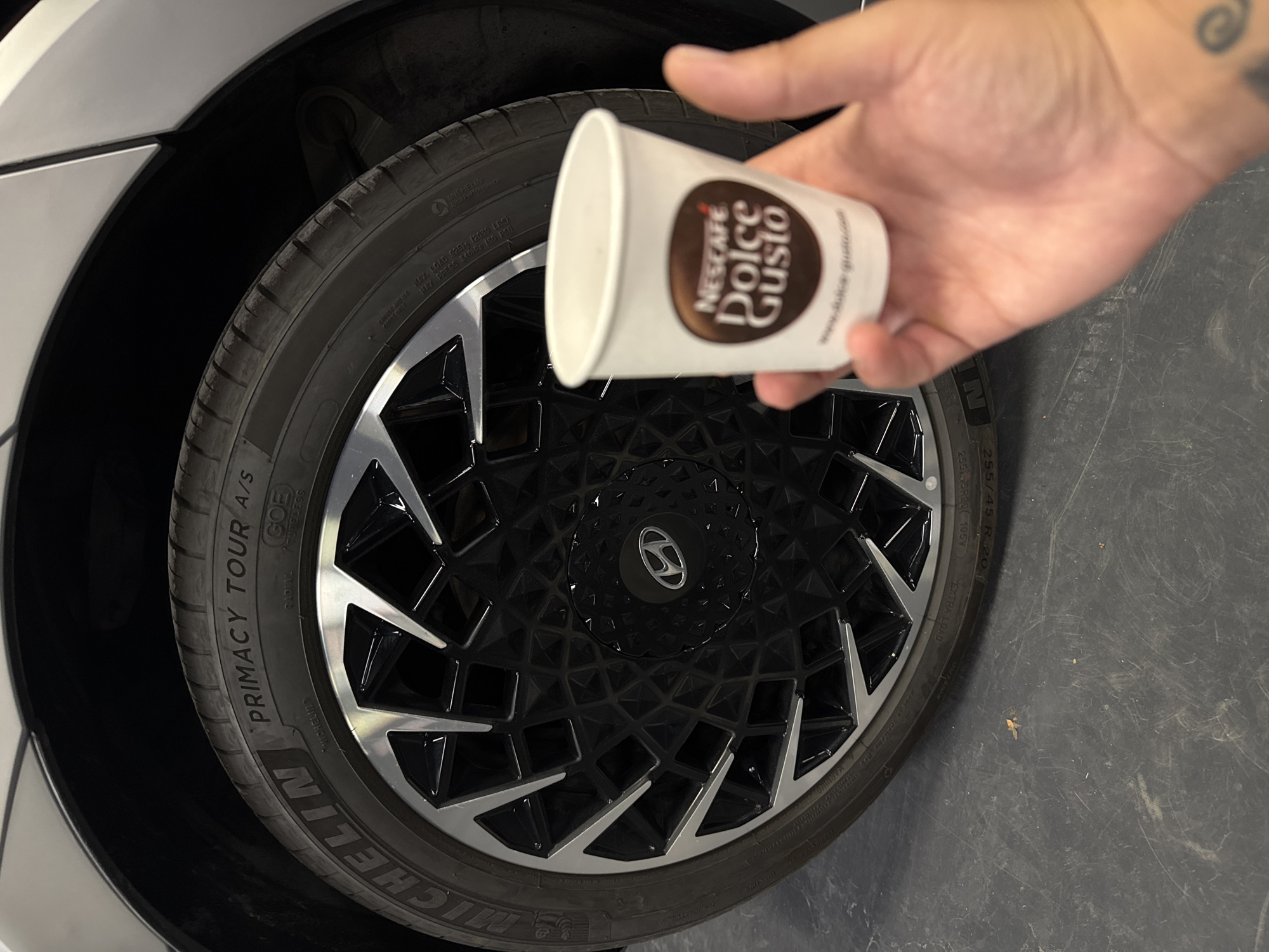 A hand, partially obscuring the lower right of the frame, holds a white paper cup with the Nescafé Dolce Gusto logo. The cup is positioned in front of a car wheel. The wheel has a black rim with a geometric, faceted design and silver accents. The tire is a Michelin Primacy Tour A/S. The background shows the dark floor of what appears to be a garage or parking area. The image does not contain enough information to determine a specific location within the city of Ruse, Bulgaria.
