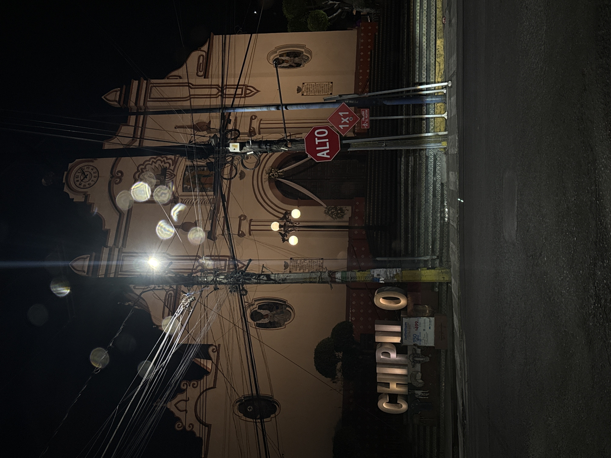 The image shows a vertical view of a street scene in Chipilo, Mexico, at night. A church with ornate white and brown trim dominates the left side of the image. An "Alto" (Stop) sign and a "1X1" sign are visible on a pole in front of the church. Numerous electrical wires are strung across the foreground. On the right, a sidewalk and street are visible. The word "CHIPILO" is spelled out in large, illuminated block letters on the sidewalk.