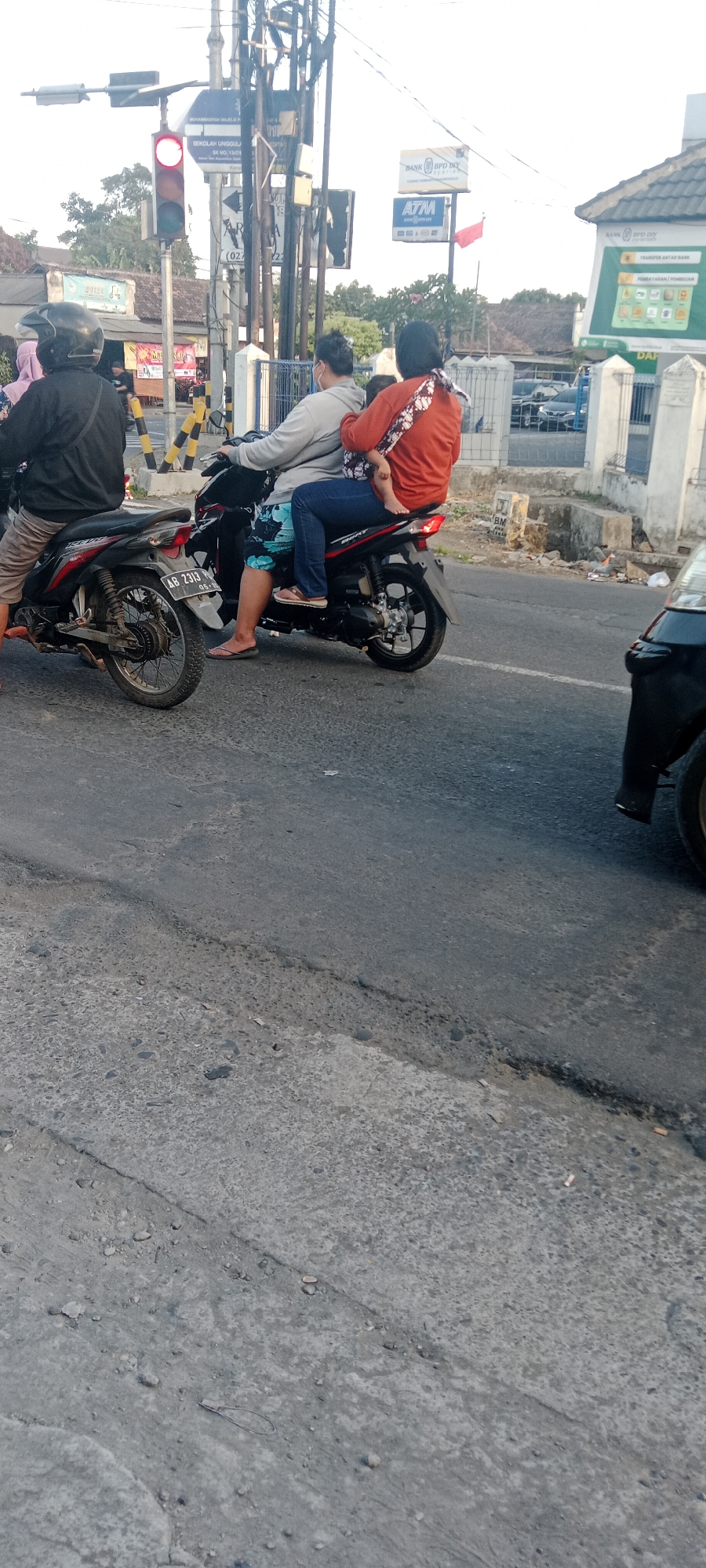 The image depicts a street scene in Maguwoharjo, Indonesia, likely near a traffic light intersection. In the foreground, two motorcycles are stopped at a red traffic light. The motorcycle on the left carries a person wearing a black jacket and a helmet. The motorcycle on the right has three occupants: the driver, a passenger holding a baby on their lap, and another passenger behind the driver. The passenger holding the baby is wearing an orange shirt and blue jeans, and the baby is wearing a patterned cloth. The driver of this motorcycle is wearing a grey hoodie and patterned shorts. The road appears to be paved with asphalt and shows signs of wear. In the background, there are various signs, including a traffic light, a sign for "BANK BPD DIY", and an "ATM" sign. Buildings are also visible behind the signs. The overall atmosphere suggests a typical urban environment during daylight hours.