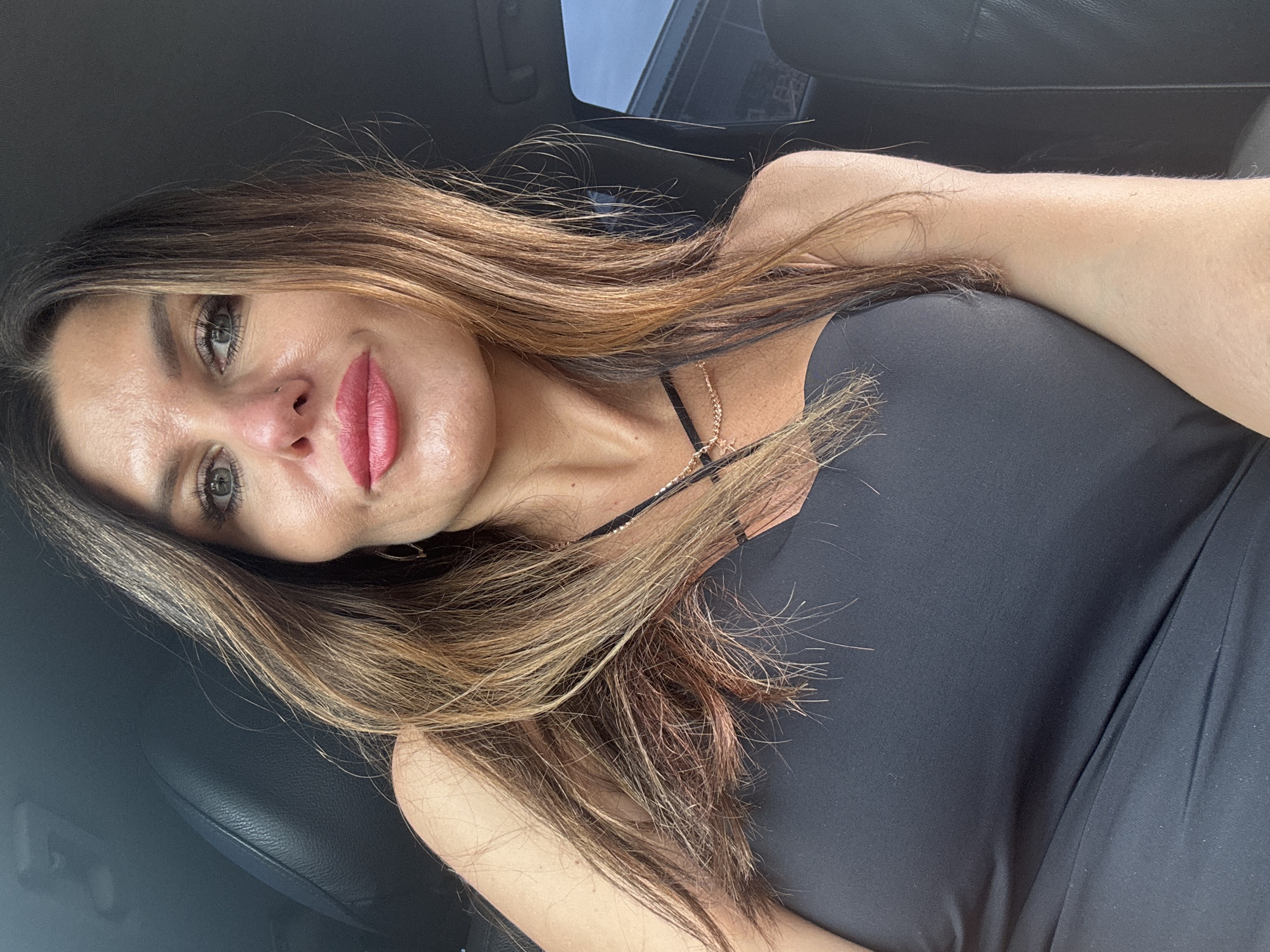 A selfie-style portrait shows a woman inside what appears to be a car, likely in Chaikovsky, Russia, given the context. The woman is positioned to the left of the frame, looking directly towards the camera with a slight, subtle smile. She has long, multi-toned hair, primarily brown with lighter blonde highlights, which cascades over her shoulders and chest. Her eyes are a striking gray-green, and she is wearing makeup, including prominent pink lipstick. A small nose piercing is visible on her right nostril.
She is wearing a dark, possibly black, strapless or spaghetti-strap top. A delicate golden necklace with a small pendant is visible around her neck. The background consists of the car's interior, featuring dark leather or fabric upholstery on the seats and what looks like a dashboard or roofline towards the top and right, suggesting daylight conditions outside. Her right arm is visible in the lower right portion of the frame. No visible text is present. The overall mood of the image is casual and relaxed.