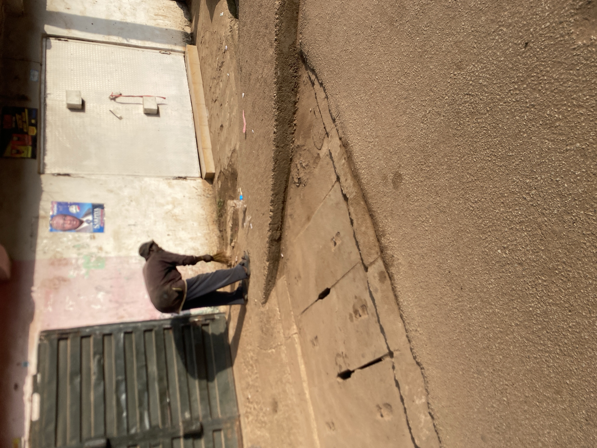 An individual is sweeping the ground in front of a building with a metal corrugated door and a large, white metal door on the right. A poster of a man's face is visible on the wall. The ground is paved with concrete and asphalt, with a manhole cover present. There is some debris on the ground, including a blue plastic bottle and pink paper. The scene appears to be an outdoor commercial area in Bukuru, Nigeria.