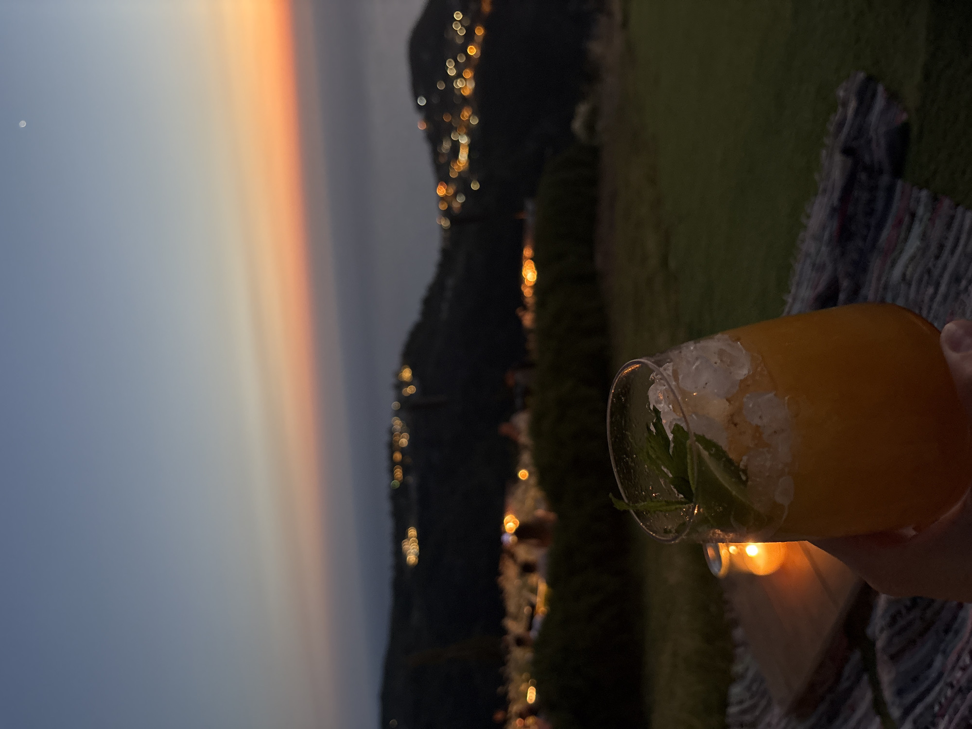 A hand holds a clear glass filled with an orange beverage and ice, garnished with mint, with a blurred background of a dusky sky and distant city lights on rolling hills. The scene appears to be set outdoors at dusk, possibly on a picnic blanket, with a lit candle visible on a wooden surface. The horizon shows a gradient of blues and oranges, transitioning into the dark silhouettes of hills dotted with illuminated structures. The Municipal Unit of Sfakiates, Greece, is known for its mountainous terrain and traditional villages, which aligns with the visual elements of the image.