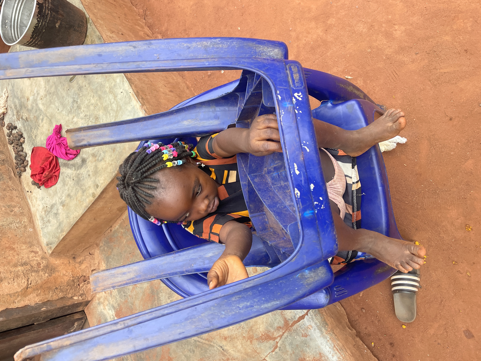 A young person with dark skin is angled upward inside a blue plastic chair. The person has braided hair with colorful beads. A hand reaches from the chair's left side, and feet extend from the chair's right side. One foot has a gray and white sandal next to it. The chair is on a reddish-brown surface that may be dirt or brick. To the left, there is a concrete or stone surface with a bucket, some nut-like objects, and red and pink cloths. The image appears to be taken in the city of Ogbe, Nigeria.