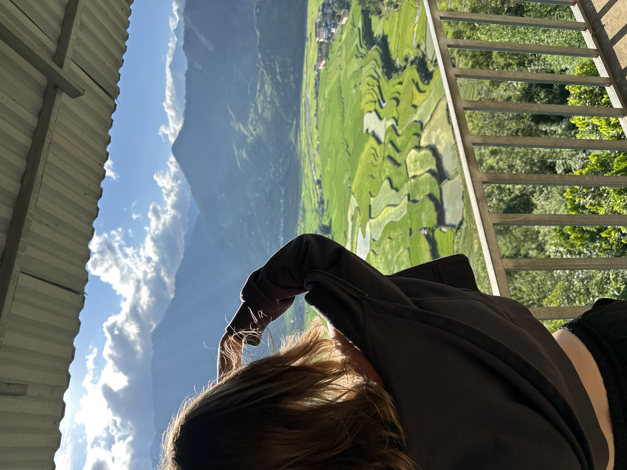 The image depicts a young woman, viewed from behind, standing on a balcony or elevated platform, gazing out at a mountainous landscape with terraced rice paddies. The scene is located in Tú Lệ, Vietnam.
In the foreground, a corrugated metal roof extends across the top of the frame, partially obscuring the sky. Below this, a silver-colored metal railing runs horizontally across the lower mid-section of the image, defining the viewing area. The woman, with long, wavy brown hair, is silhouetted against the bright landscape. She wears a dark grey or black long-sleeved top, possibly a hoodie, and dark pants. Her right arm is raised, with her hand near her forehead, possibly shielding her eyes from the sun or adjusting her hair. Sunlight catches the edges of her hair and the fabric of her sleeve, creating a halo effect.
The midground reveals an expansive valley filled with vibrant green rice terraces, some reflecting water. A winding river or stream traverses the valley floor. Several buildings, indicative of a small town or village, are visible nestled amongst the fields and at the base of the mountains.
In the background, multiple layers of verdant mountains rise under a bright blue sky. Fluffy white clouds are scattered across the sky, with visible sun rays piercing through, creating patches of light and shadow on the distant slopes. The overall impression is a scenic, sunlit view over a rural Vietnamese landscape.