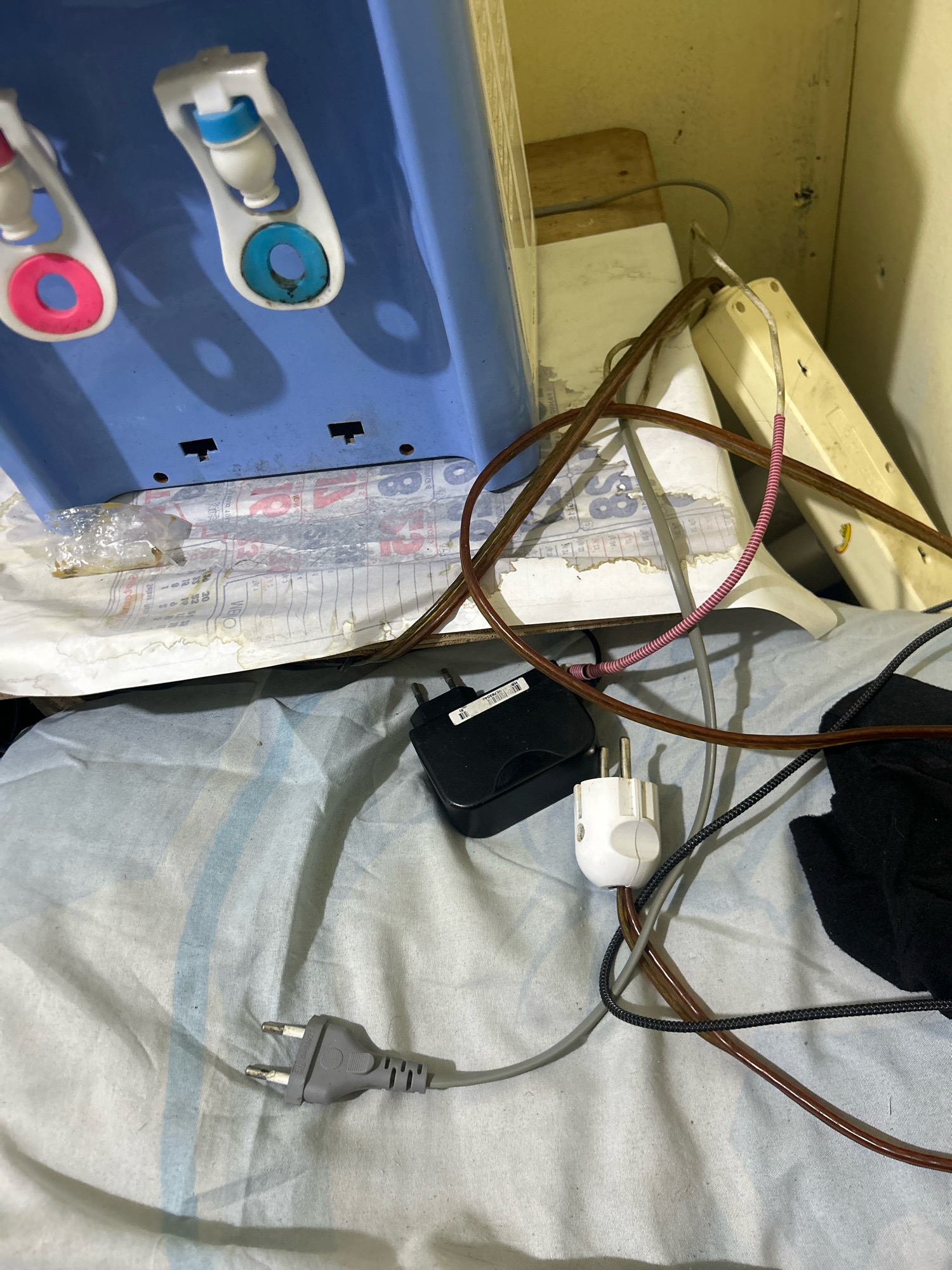 A blue water dispenser is positioned on a surface covered with a patterned cloth. Two spigots are visible on the dispenser, one with a pink ring and the other with a blue ring. Below the dispenser, a piece of paper with printed text, possibly a newspaper, is partially visible. Several electrical cords and plugs are scattered around the base of the dispenser. A black power adapter, a white plug with a European-style adapter, and a grey plug with two prongs are among the items. A beige power strip is also visible in the background. The overall scene appears to be indoors, with a muted color palette and a slightly cluttered arrangement of objects. The location is identified as Sukanagara, Indonesia.