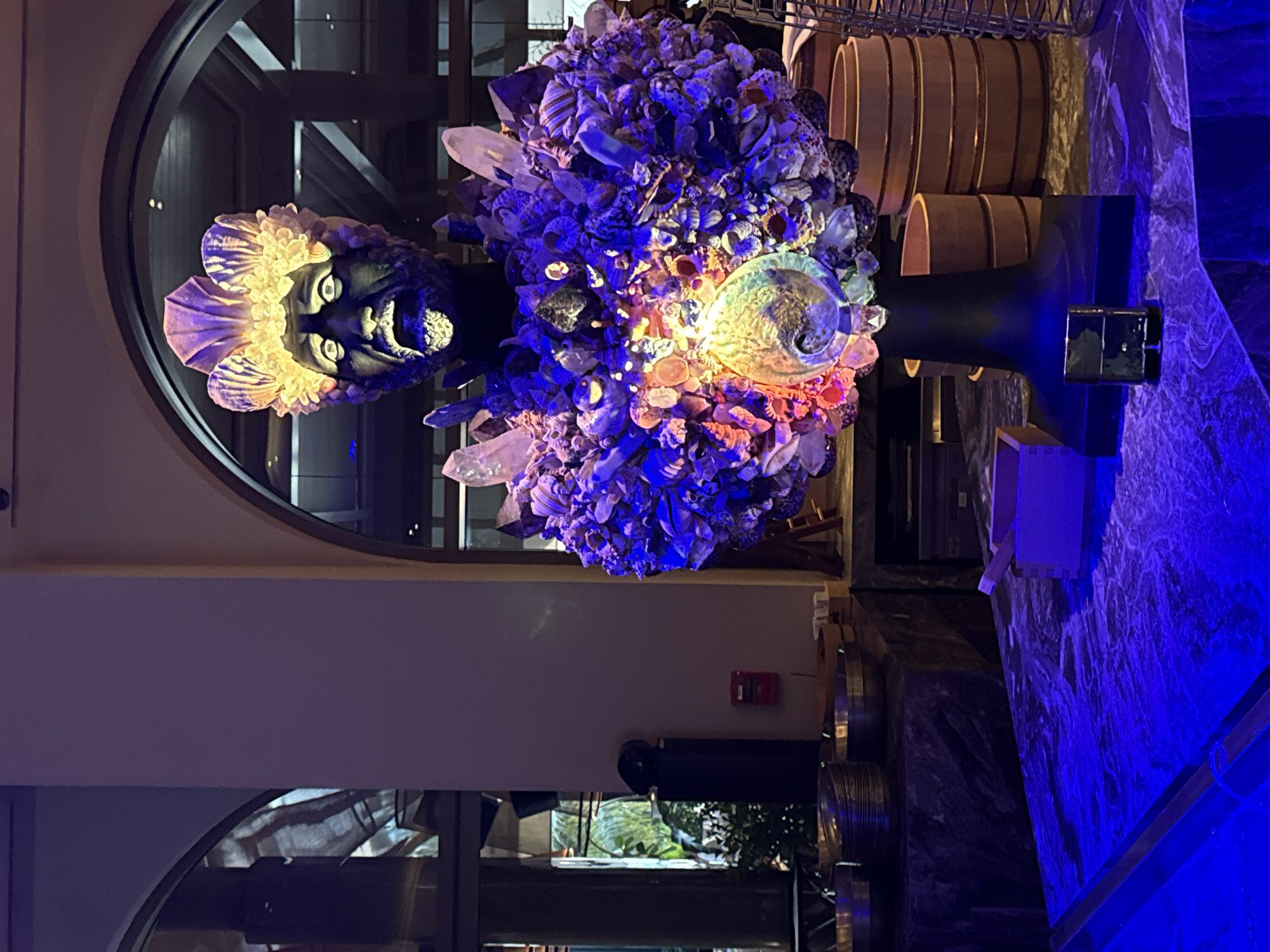An elaborate sculpture of a bust with an open mouth and wide eyes is displayed on a dark stand in Dubai, United Arab Emirates. The sculpture is adorned with a crown of shells and a densely packed sphere of seashells, crystals, and other natural elements. The sphere is illuminated with vibrant blue and yellow lights, creating a dramatic effect. In the foreground, a dark marble surface is bathed in blue light, with a small metallic object, possibly a lighter, placed on it. In the background, arched windows reveal glimpses of an indoor environment, possibly a hotel or restaurant, with tables and decorative elements visible. The overall scene suggests an artistic installation in a public or commercial space.