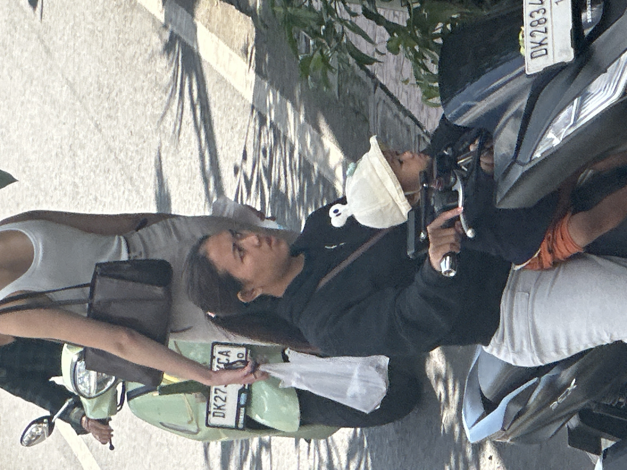 A young Indonesian woman, wearing a black hoodie and light-colored pants, is riding a black scooter. She has her hair pulled back in a ponytail. A young child, wearing an orange garment and a white hat with two white pom-poms, is seated in front of her on the scooter. The child is looking to the right. A second woman, partially visible, is standing next to the scooter on the left, holding a dark brown purse and a white plastic bag. The scooter has a green front fairing and a license plate that reads "DK27XCA". The scooter is parked on an asphalt road, with shadows from trees cast on the surface. The scene is set in the city of Ubud District, Indonesia.