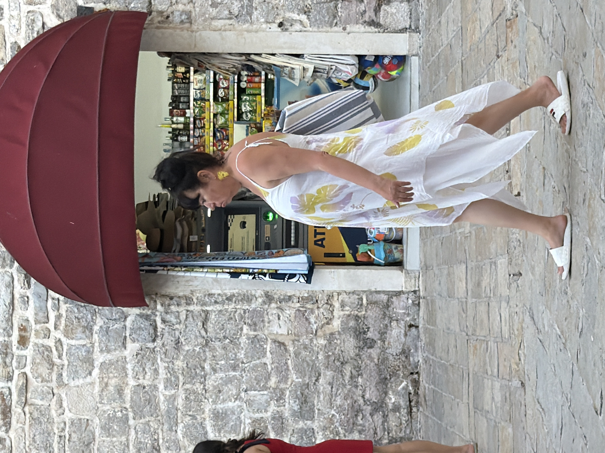 A woman with dark hair, wearing a white dress with a floral pattern, walks past an open shop. She has yellow earrings and is looking down. Her dress has spaghetti straps. She is wearing white flip-flops. In the background, the shop is stocked with various items, including bottles and snacks. There's a red awning extending from the shop. The scene is set against a backdrop of a stone building and a cobblestone path. The lighting suggests it is daytime. Further down the path, another person is partially visible, wearing a red dress. The overall impression is of a sunny day in a Mediterranean town. The text "ATI" is visible on a yellow sign in the shop.