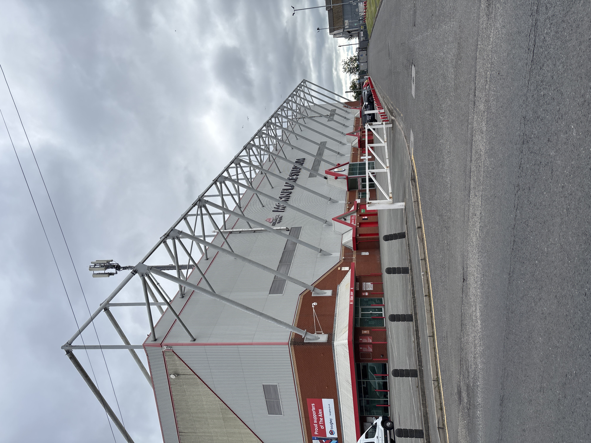This is a full-shot, eye-level, outdoor, daytime photograph of Mornflake Stadium, located in Crewe, United Kingdom. The stadium, also known as Gresty Road, is a football stadium with a predominantly red and white exterior. A portion of the grandstand, featuring a metal support structure and grey cladding, dominates the left side of the frame. Red trim lines the upper edge of the building and the roofline.
In front of the stadium, a paved area serves as a parking lot. A white van is parked on the left, and several cars are lined up further down the road on the right. A white barrier gate stands across the entrance to the parking area. Black bollards line the edge of the paved area closest to the stadium. The sky is overcast and grey, with a few scattered birds visible. A cell tower is mounted on the stadium's roof. A banner on the left side of the building reads "Proud supporters of The Alex."