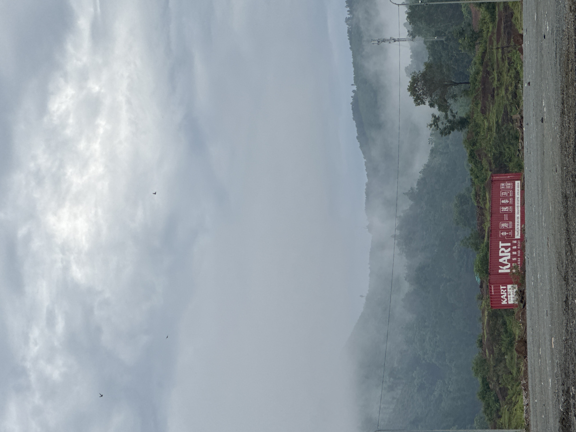 The image depicts a misty landscape with rolling hills covered in dense green foliage. The sky is overcast with dramatic grey clouds. In the foreground, there are two red shipping containers, each with white lettering. The left container displays "KART" and some text in what appears to be Asian characters, along with a barcode. The right container also prominently features "KART" and additional Asian characters, including "中", and further text and a barcode. To the right of the containers, a large cell tower stands against the backdrop of the misty hills. The ground in the immediate foreground is a rough, gravelly surface. The scene suggests an outdoor storage or logistics area, likely on the outskirts of a town or in a more rural setting, given the natural landscape dominating the background.