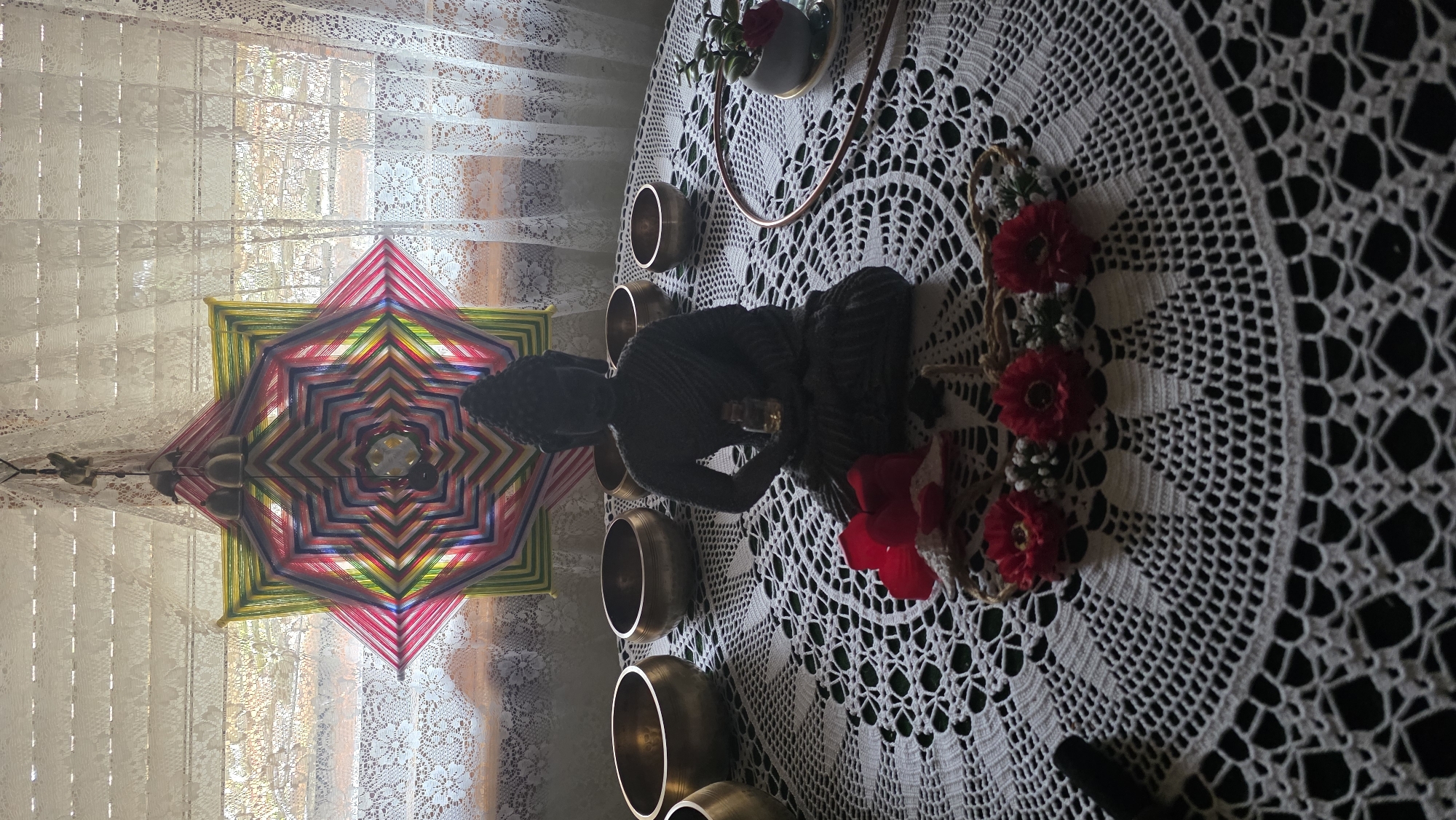 An interior shot depicts a serene, spiritually-themed arrangement. The central subject is a dark-colored statue of Buddha, seated in a meditative pose, placed on a white, intricately crocheted or lace tablecloth. Around the Buddha are several small, brass-colored singing bowls. To the right of the statue, on the tablecloth, are decorative elements including a string of red artificial flowers or berries, and a small potted plant with a red floral accent, alongside a copper-colored ring.
In the background, positioned to the left of the Buddha statue, hangs a large, star-shaped yarn mandala (often known as a God's Eye). This colorful piece features concentric layers of red, pink, blue, green, and yellow threads, creating a vibrant geometric pattern. Behind the mandala and extending across the entire background are white lace curtains covering a window, through which soft, natural daylight filters. The sunlight creates subtle shadows and highlights on the lace, indicating it is daytime. The overall ambiance is peaceful and contemplative. No specific activity is taking place, but the setup suggests a space for meditation or spiritual reflection. There is no visible text in the image. The location context is Blagoevgrad, Bulgaria.