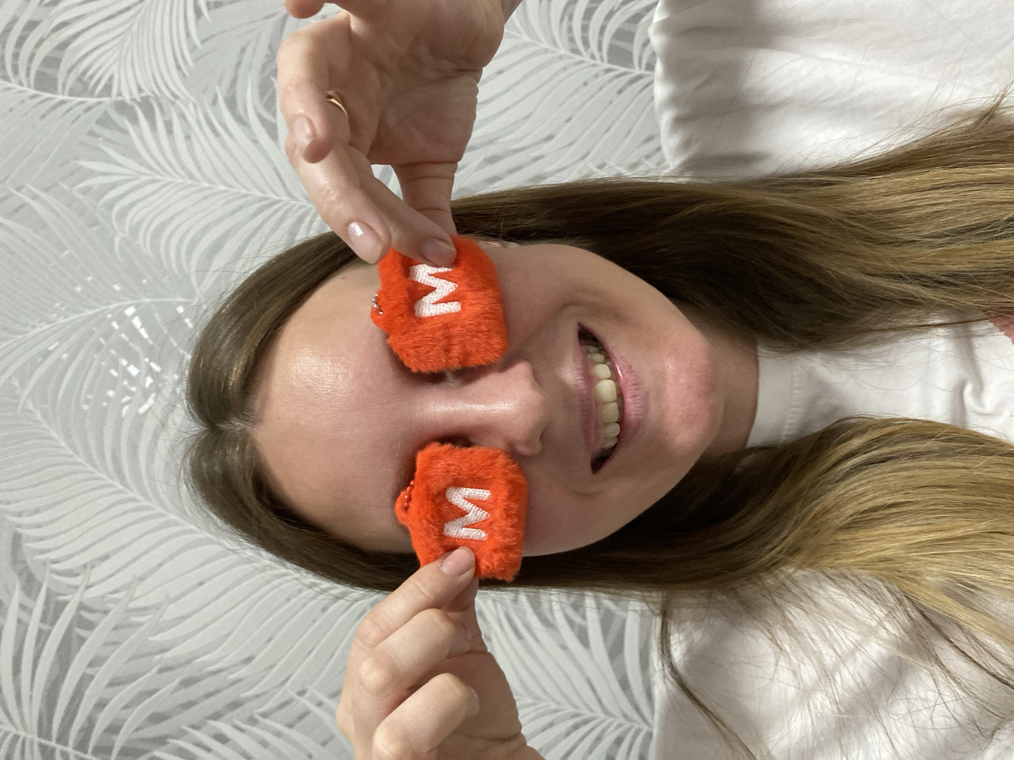 A woman is holding two orange, furry objects with a white "M" on them over her eyes, smiling and looking at the camera. Her hair is brown and long, and she is wearing a white shirt. She has a ring on her finger. The background is a gray and white leaf pattern. There is no evidence to suggest a specific location in Noviy, Russia.