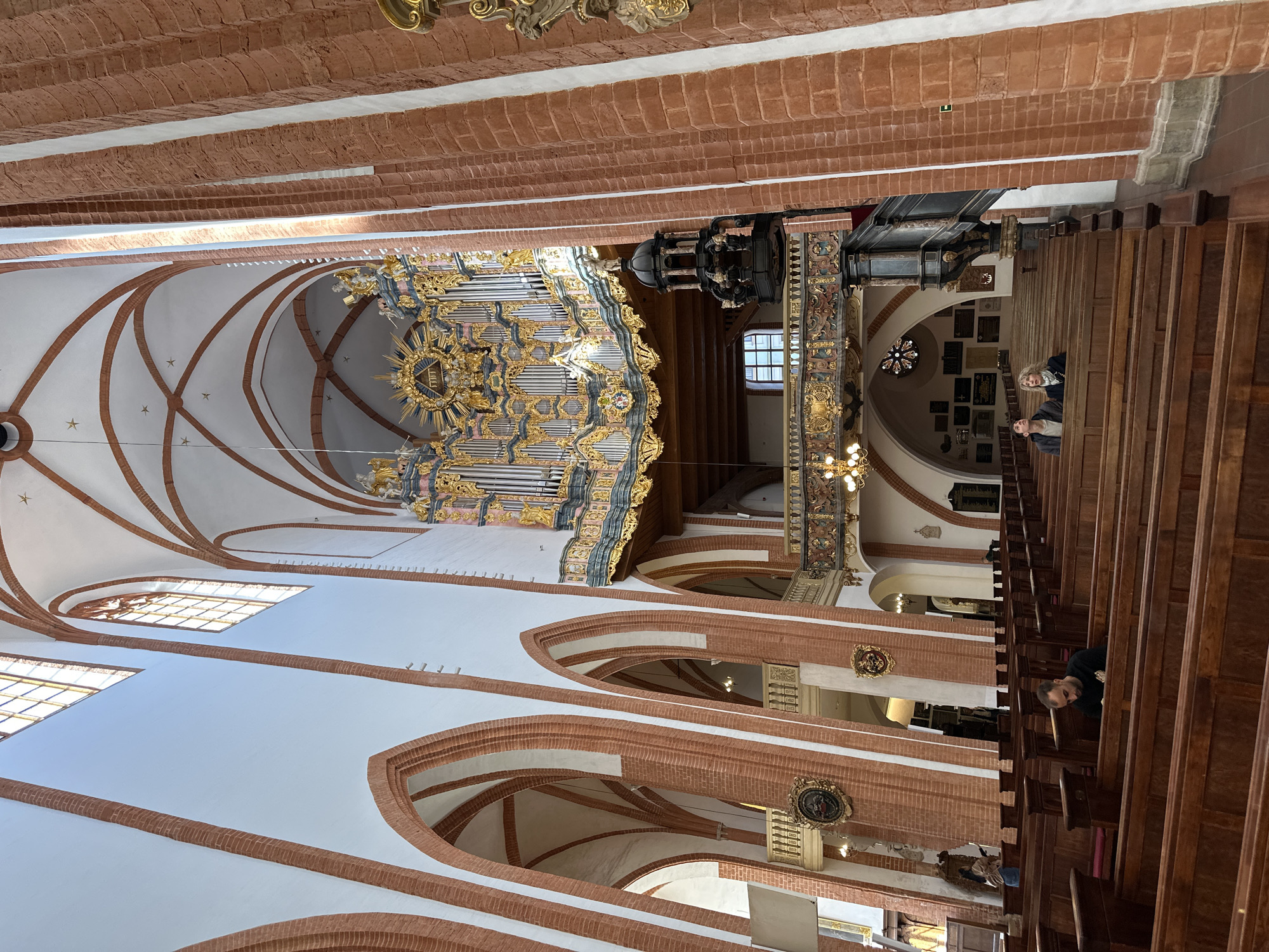 The image captures the interior of a church, likely located in Wrocław, Poland, based on its architectural style. The camera angle is slightly upward, showcasing the high, vaulted ceiling with brick arches and white plaster infills. A large, ornate pipe organ dominates the upper left quadrant of the frame. It is intricately decorated with gold and blue accents.
To the right of the organ, a pulpit is visible, positioned above the rows of wooden pews. People are seated in the pews, including two individuals closer to the back and one individual more forward.
The lighting is a mix of natural light streaming through the windows and artificial lighting from chandeliers. The color palette predominantly features warm tones from the brick and wood, contrasted by the white plaster and cooler tones in the organ’s detailing. The architecture reflects a gothic or neo-gothic style, with ribbed vaults and pointed arches.