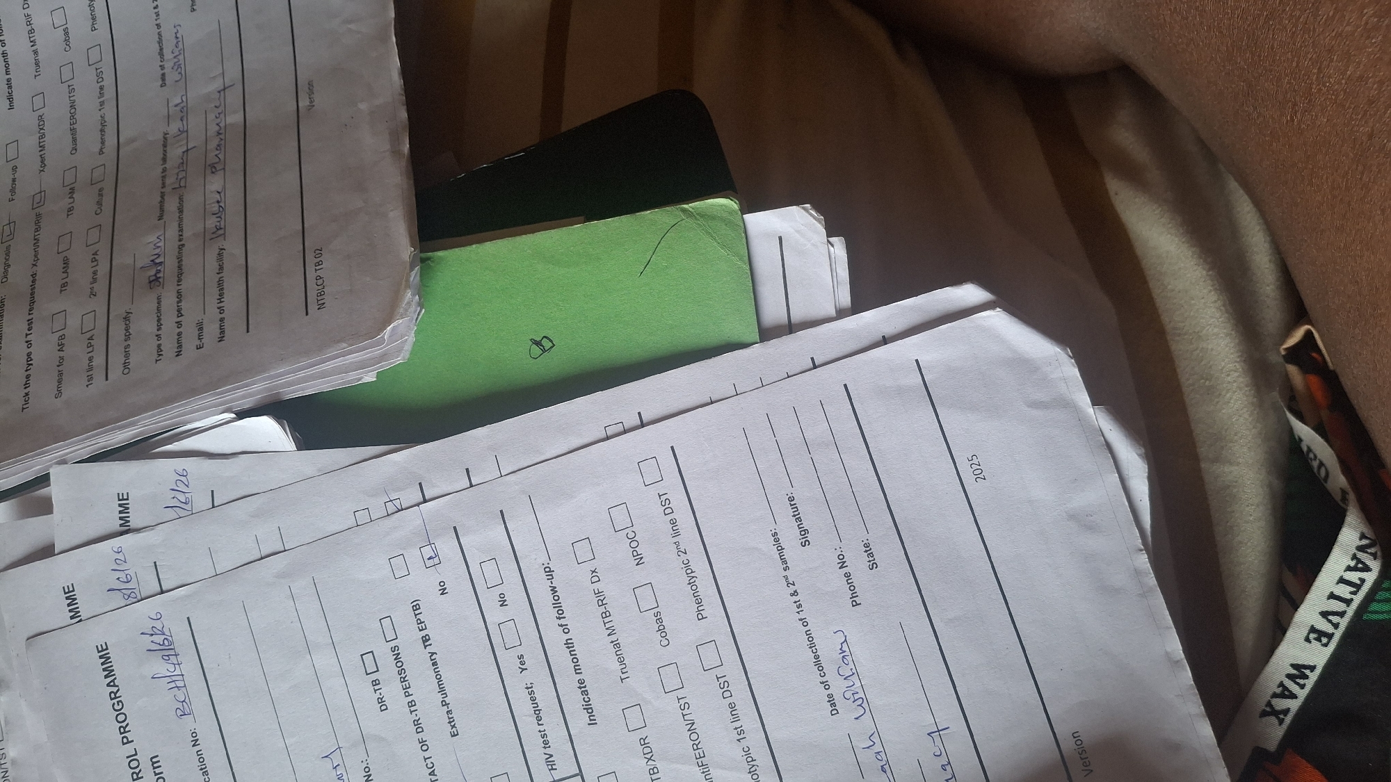 A close-up, high-angle shot depicts a stack of medical laboratory forms on a beige, textured surface that appears to be a bed or soft furnishing. The forms are primarily white with black text and lines. Some forms are partially obscured by others. One form on the left has handwritten text in blue ink, including "BCH/19/6/26" and "8/6/26". Another form has handwritten text in blue ink that appears to be a signature and name "Sarah Williams". A green folder is visible on top of some of the forms. The lighting suggests an indoor environment, possibly a bedroom, with shadows cast across the scene. The background is out of focus, showing indistinct shapes and textures. A portion of dark fabric with "NATIVE WAX" printed on it is visible at the bottom of the frame.