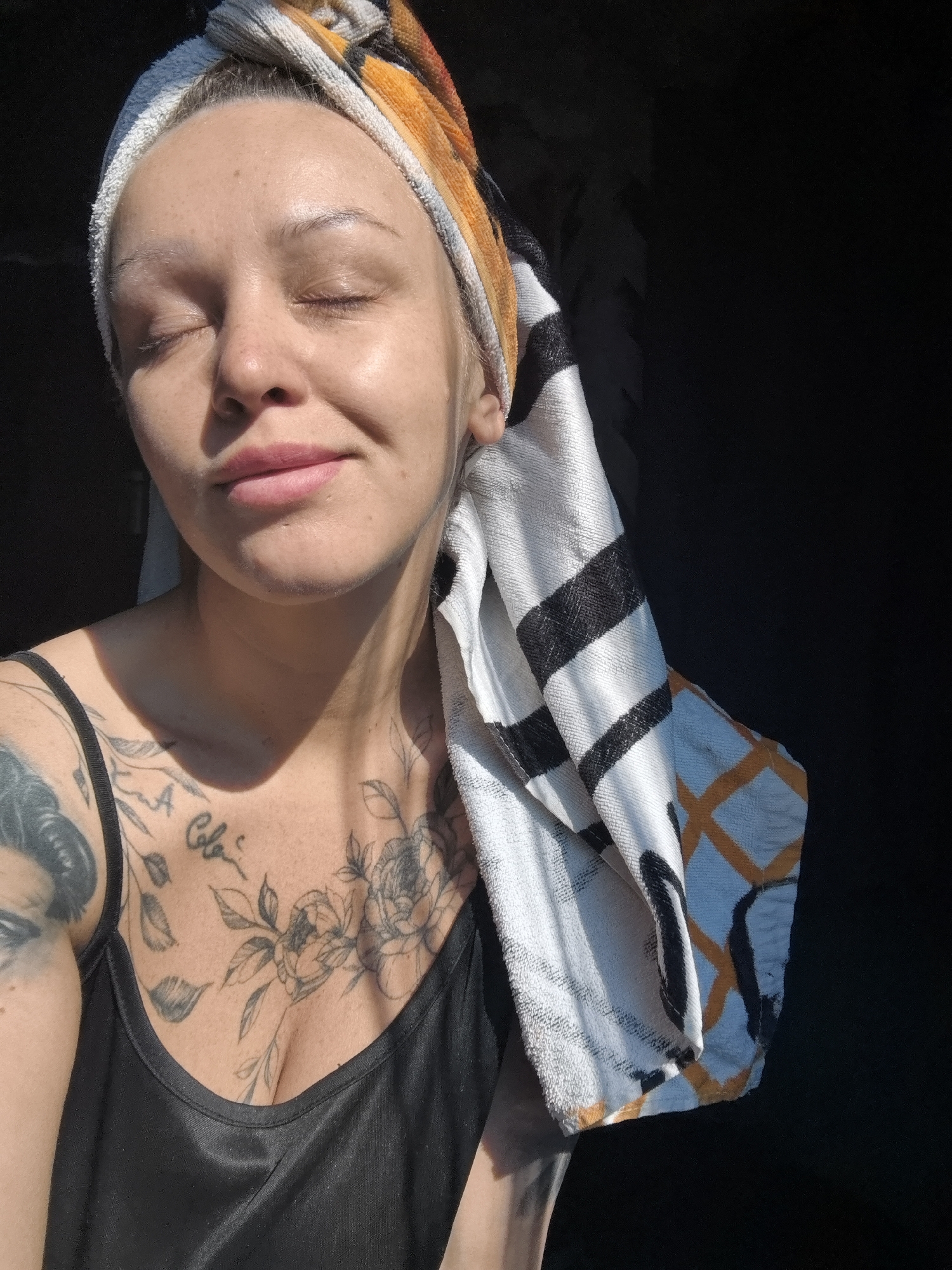A close-up, high-contrast portrait captures a person, likely female, with fair skin and closed eyes, basking in strong, direct sunlight. She has a slight, serene smile, suggesting a moment of peace and contentment. A white towel with black stripes and orange geometric patterns is wrapped around her head, indicating recent hair washing or a self-care routine.
The bright light, originating from the left, illuminates her face and upper chest, casting the right side of her head and a significant portion of the background into deep, undifferentiated shadow. The background is a stark, featureless black. She is wearing a simple black tank top or camisole.
Visible tattoos adorn her upper body. On her left shoulder, a large, dark tattoo, possibly a portrait or figure, is partially seen. Her chest and décolletage are covered with several intricate floral and leafy outline tattoos, including a prominent rose. Among these, a clear script tattoo reads "A Cole." The image conveys a sense of tranquility under the warm sun during daytime, likely indoors in Noviy, Russia, given the context.