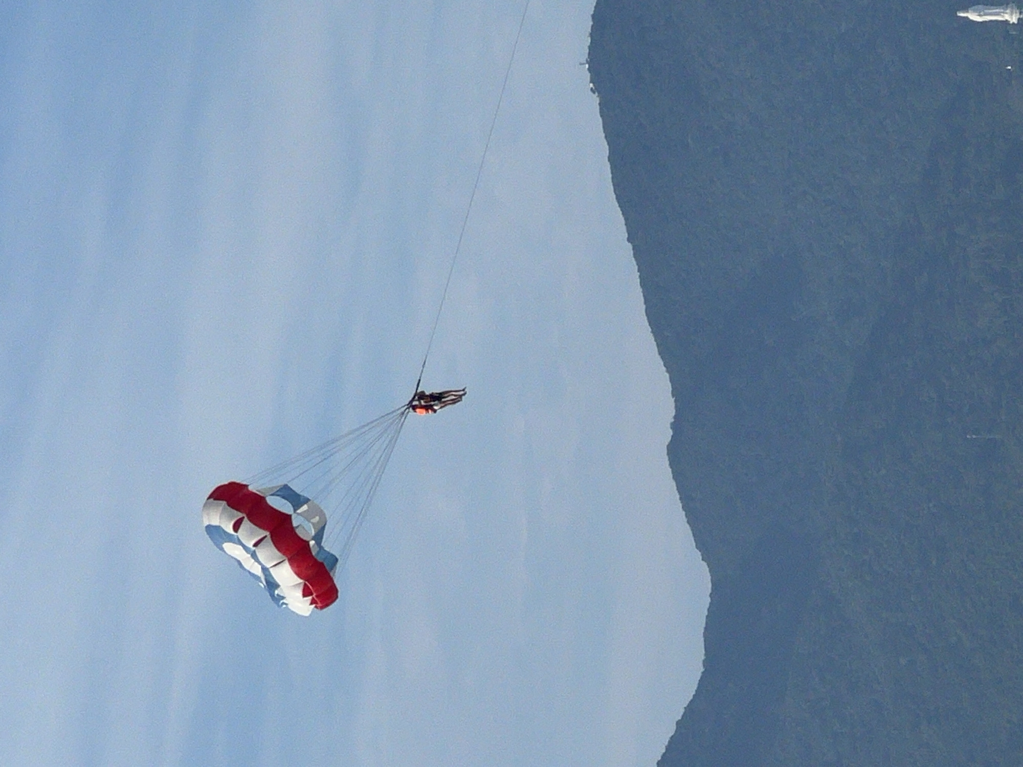 In this landscape orientation photo, two people are parasailing against a clear blue sky. One person is wearing a red and black life vest and is attached to a white, red, and blue parachute, which is partially inflated and angled downwards. The parachute is suspended by several white ropes. The second person, also in a life vest, is positioned above the parachute, trailing behind a tow rope. They appear to be in mid-air, enjoying the activity.
The background is dominated by a large, dark blue mountain range that occupies the right side of the frame, its peak catching a hint of light at the very top. The clear sky suggests it is daytime, likely with fair weather conducive to outdoor activities. The photo seems to be taken from a slightly elevated perspective, looking up towards the parasailers and down towards the mountain range.
While no text is visible, the activity of parasailing, combined with the presence of a mountainous coastal landscape, hints at a tourist destination. Given the location context of Đà Nẵng, Vietnam, the scene likely depicts a recreational activity along the coast. The overall mood is one of adventure and leisure.