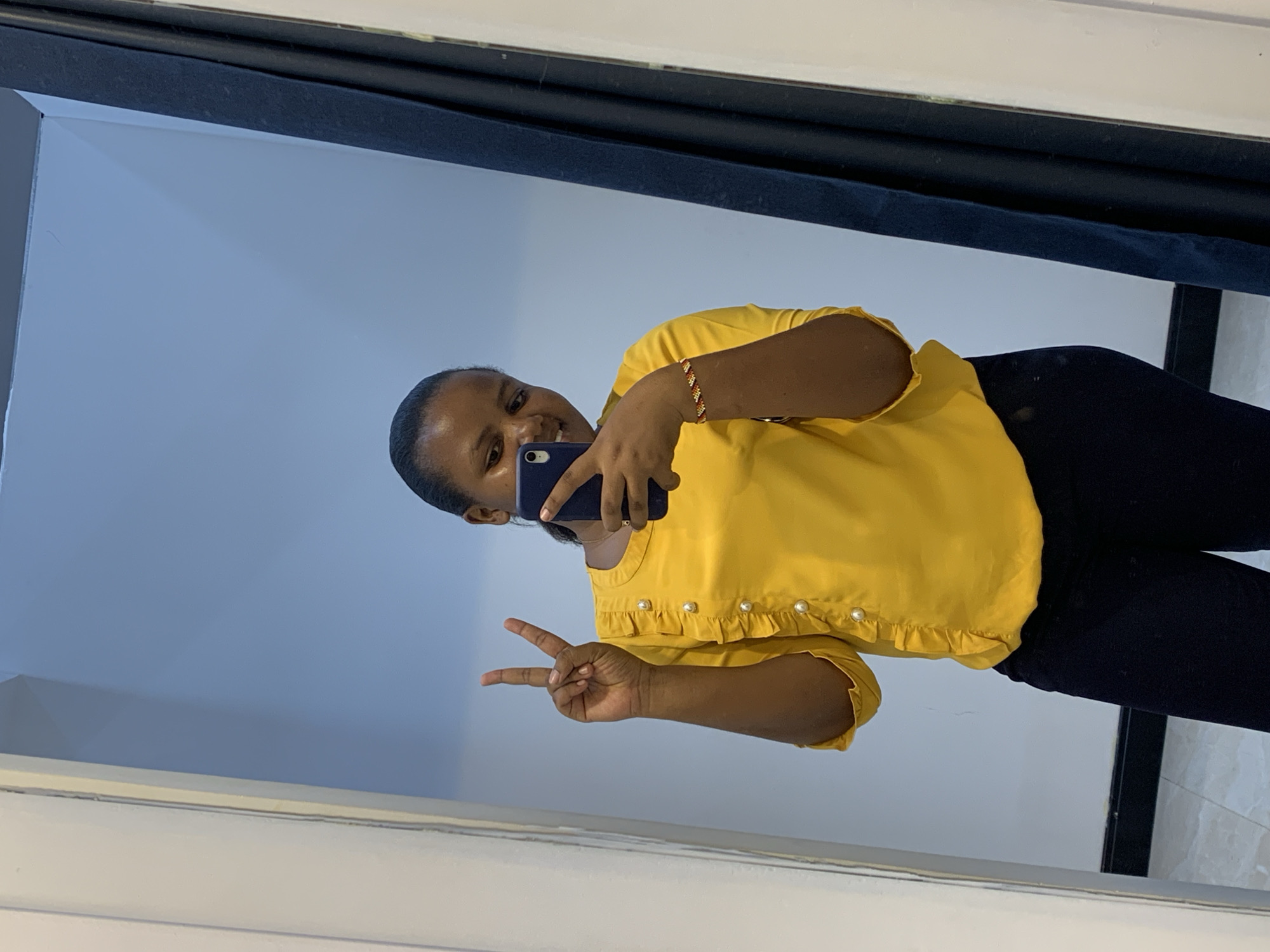 A young woman is taking a selfie in a mirror. She is wearing a yellow blouse with pearl details and black pants. She is making a peace sign with her left hand and holding a blue smartphone in her right hand. The location appears to be indoors, possibly in a room in Dar es-Salaam, Tanzania based on the subject's features and the clothing worn. The wall behind her is a light blue color. There is a dark blue draped covering above the mirror. The floor appears to be tiled.