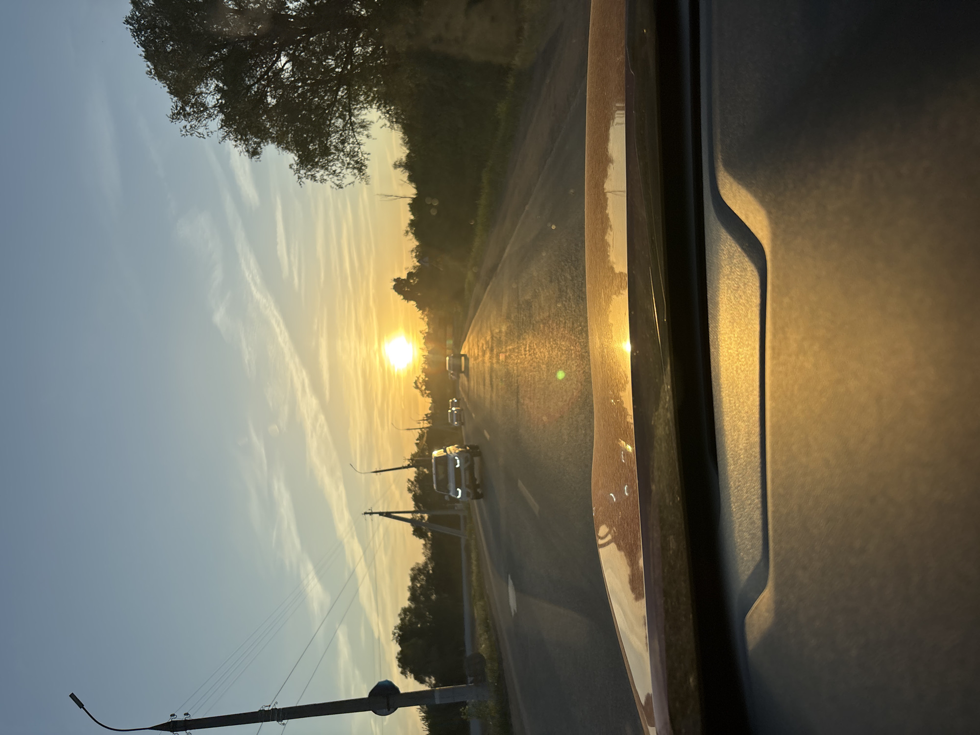 The image is taken from inside a vehicle traveling on a road, likely in the vicinity of Kabanikha, Russia, given the landscape features and the context of the request. The sun is setting directly in the line of travel, causing significant glare on the road surface and vehicle interior. Other vehicles can be seen on the road ahead. To the sides of the road, there are trees and vegetation, along with utility poles and streetlights. The sky is clear with some wispy clouds. The interior of the vehicle is partially visible, including a portion of the dashboard.