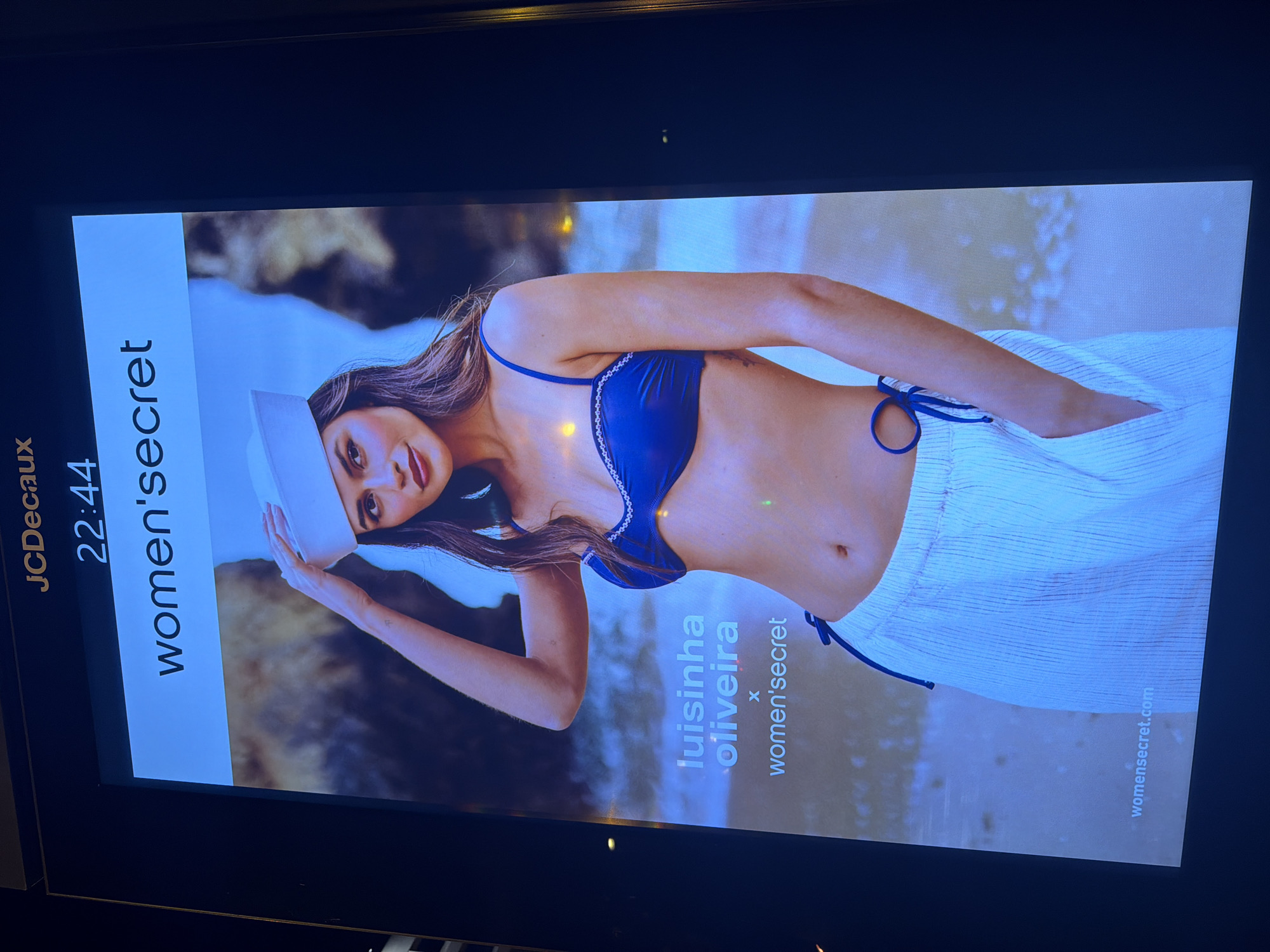 The image shows a large digital advertisement screen in Lisbon, Portugal. The screen displays a photograph of a woman wearing a blue bikini top and white striped pants, with a white sailor hat on her head. The advertisement includes text that reads "women'secret", "luisinha oliveira x women'secret", and "womensecret.com". Above the advertisement, "JCDeaux" and the time "22:44" are visible. The background of the advertisement is a blurred image of a beach scene.