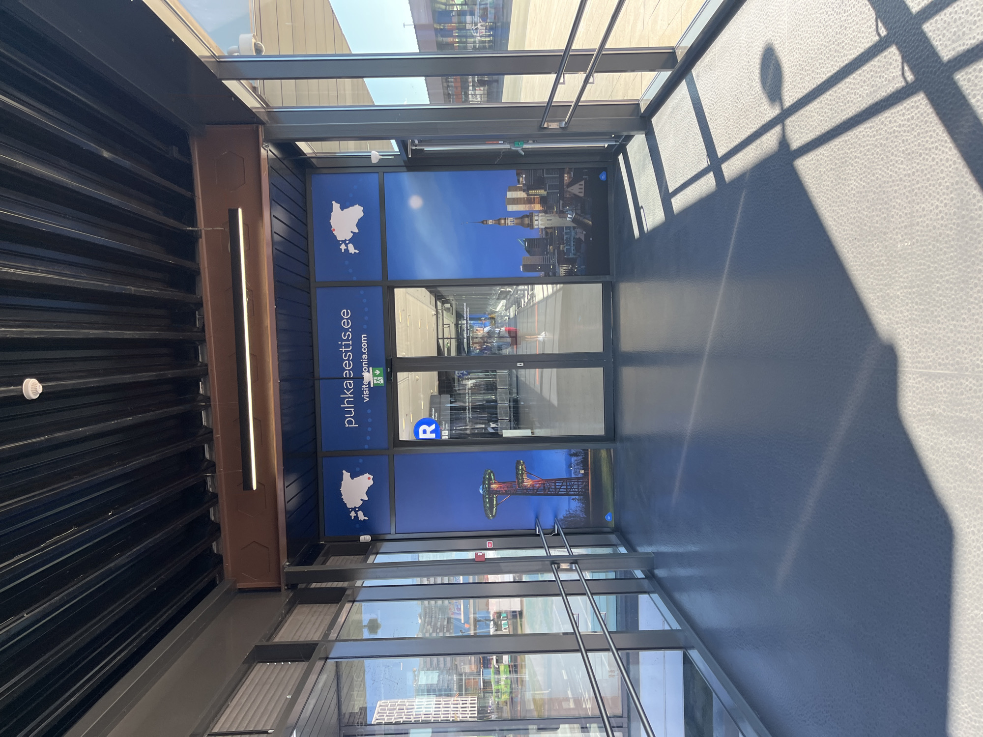 The image shows an indoor walkway with a glass facade on the right, looking out onto a sunlit outdoor area. The walkway is covered by a dark, corrugated ceiling and has a gray, textured floor. On the left, a glass wall with a view of buildings and vehicles is visible. The main feature of the walkway is a large mural depicting scenes of Estonia. The mural features a map of Estonia with a red marker, the text "puhkaeestis.ee" and "visitestonia.com", an image of a tall, illuminated tower structure, and a cityscape with a prominent spire. Double glass doors are at the end of the walkway, leading to an interior space with escalators or moving walkways. A circular blue sign with the letter "R" is visible on the door. The overall scene suggests a modern, well-lit public space, possibly an airport, train station, or convention center in Tallinn, Estonia, given the prominent depiction of the country and its capital's skyline.