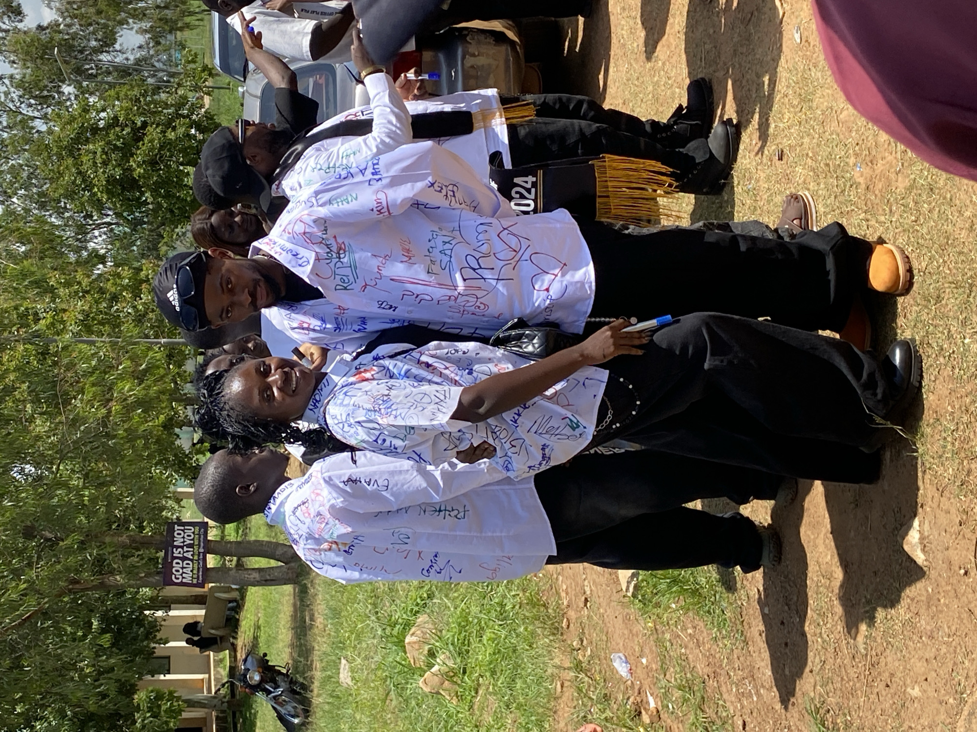 A group of young adults, likely students, are gathered outdoors in what appears to be a university or school campus setting in Durumi, Nigeria. The central figures, a male and a female, are prominently positioned and smiling at the camera. They are wearing white shirts that have been autographed and decorated with colorful markings, suggesting a celebratory occasion such as graduation. The male wears a black Adidas cap and sunglasses, and the female carries a black handbag.
Several other individuals are visible in the background, also in white attire, and some are wearing academic regalia, including a black sash with gold fringe and the inscription "2024". The environment features trees, grassy areas, and dirt pathways. In the background, a motorcycle is parked, and a car is partially visible, along with buildings that suggest an institutional setting. A sign attached to a tree reads "GOD IS NOT MAD AT YOU, HE IS MERELY IN LOVE WITH YOU." The overall atmosphere is one of joy and celebration.