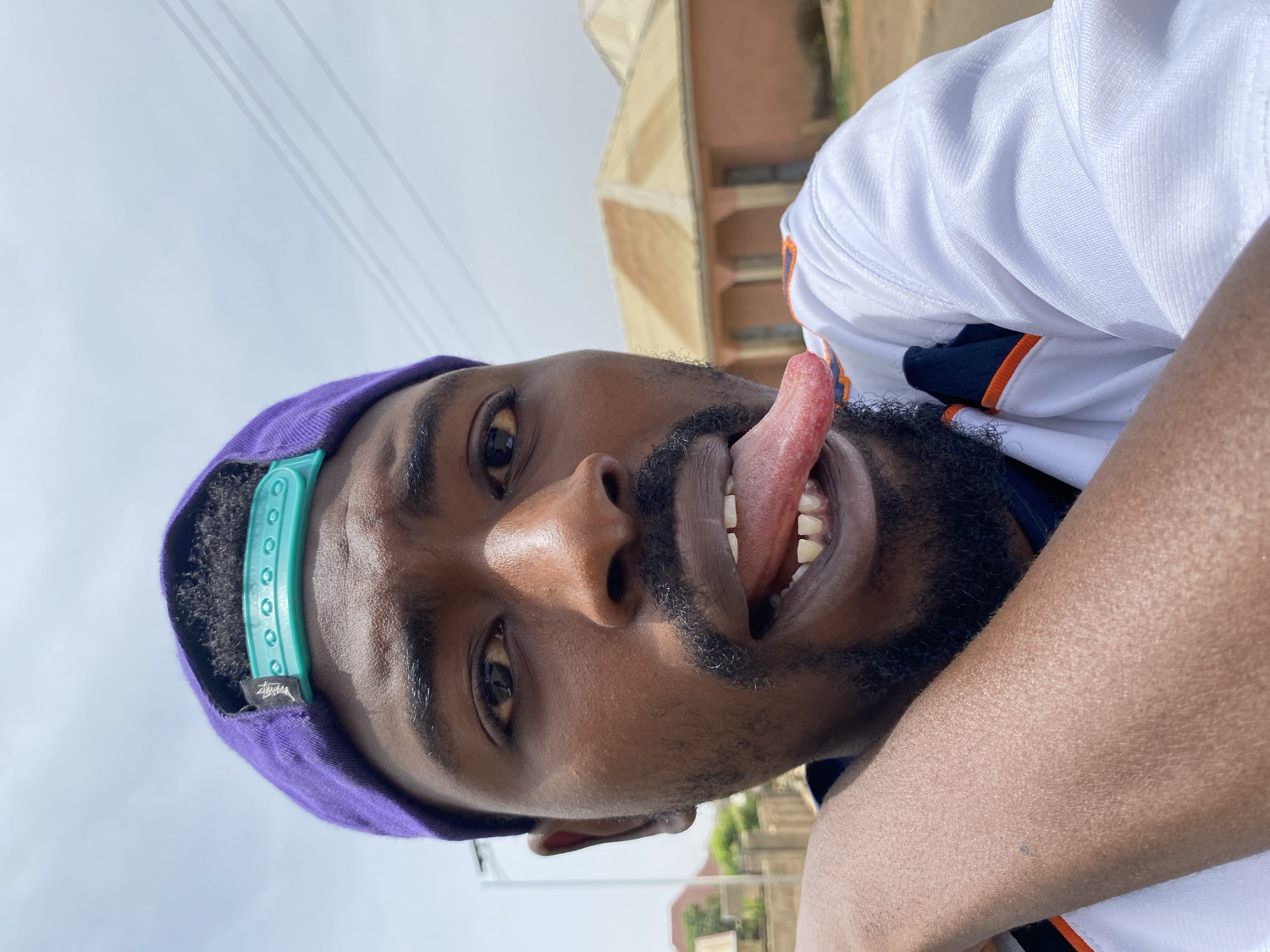 A close-up, vertically oriented selfie captures a young Black man from a low angle, looking upwards. He has a full beard and mustache and is making a playful expression by sticking his tongue out of his mouth, revealing his teeth. He wears a purple baseball cap with a prominent teal/light blue adjustable snapback, which features a small black tag with "Stussy" written in white. He is also wearing a white sports jersey, with hints of orange and navy blue visible on the collar or sleeves.
The bright, clear sky in the background indicates that the photo was taken outdoors during daytime, likely under ample sunlight. Faint architectural elements, including reddish-brown roofs and structures resembling utility lines, are visible in the distance, suggesting an urban or semi-urban setting. The image conveys a candid and lighthearted moment in Jos, Nigeria.