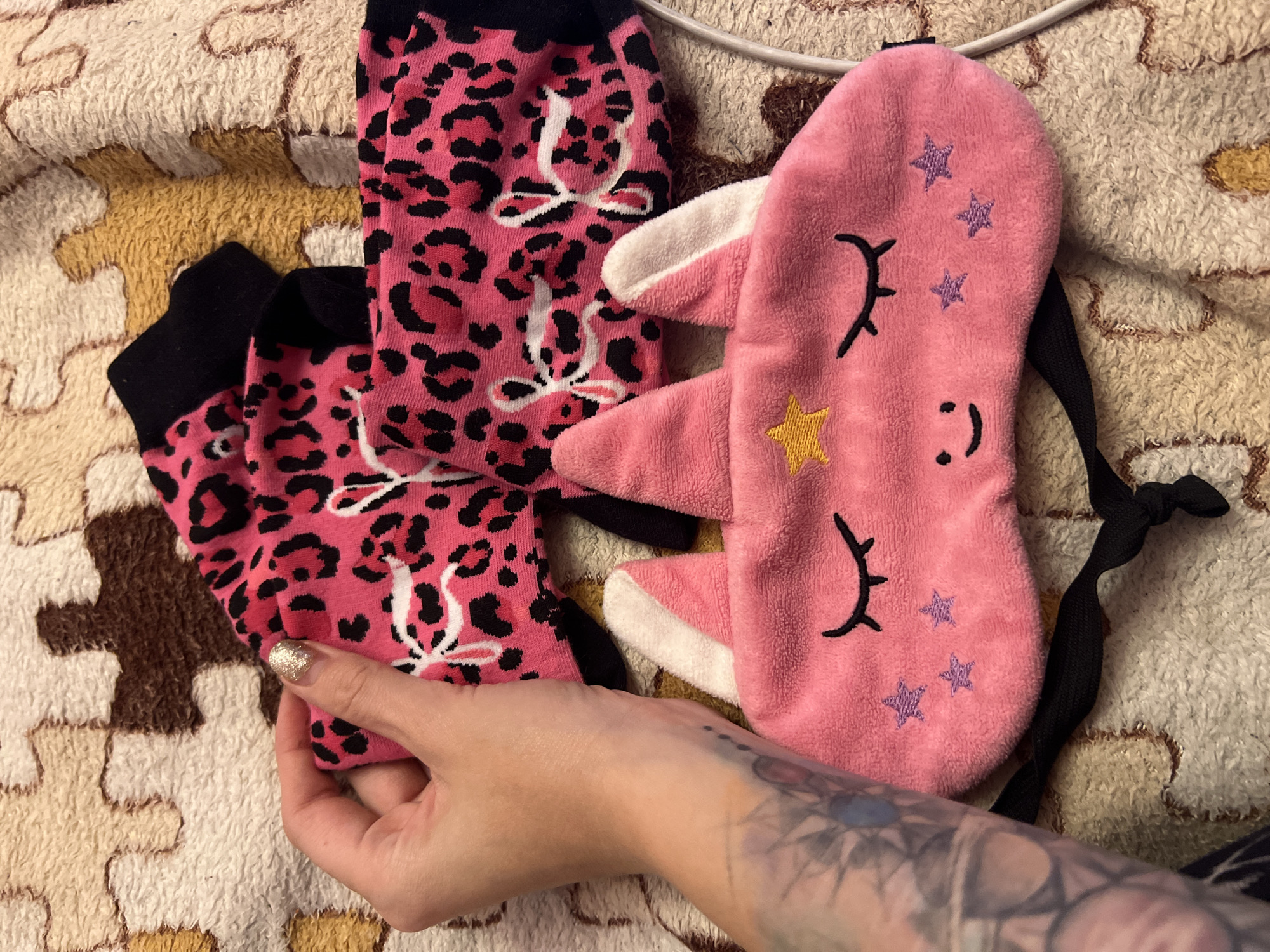 A hand with a sparkly fingernail and a visible tattoo on the wrist is holding up a pair of pink leopard-print socks. The socks have black cuffs and white bows. To the right of the socks is a pink sleep mask with a white bunny-like ear and embroidered black eyelashes, a yellow star, and purple stars. The items are laid out on a textured, fuzzy blanket with a puzzle-piece pattern in shades of cream, tan, and brown. The overall impression is one of comfort and playful, possibly feminine, items. The setting appears to be indoors, as suggested by the cozy blanket and the typical placement of such items. There is no indication of time of day or weather. No visible text.