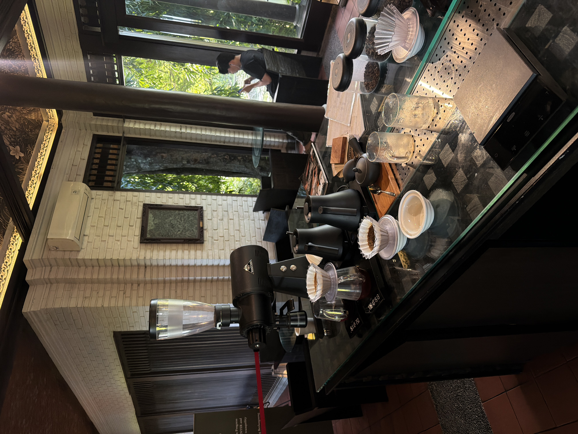 The image shows a coffee preparation station inside a building, potentially a cafe, in Sukawati, Indonesia. A black coffee grinder is prominently positioned on a black counter with a glass top. Other coffee-related items like coffee beans, filters, cups, and brewing equipment are arranged nearby. A person is visible in the background, standing by a window and looking at a mobile phone. The room has brick walls and dark wooden beams.