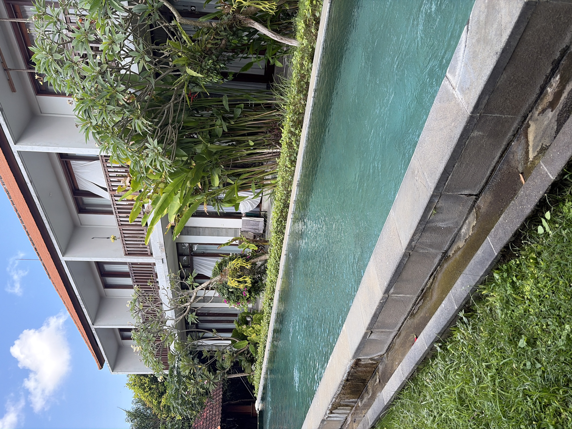 The image shows a long, rectangular swimming pool with turquoise water, situated outdoors. The pool is bordered by a stone coping and surrounded by lush green grass and tropical vegetation. Behind the pool is a multi-story building with a red-tiled roof, white walls, and balconies. The building appears to be a hotel or resort. The architecture suggests a tropical or Asian setting. The sky above is blue with some white clouds. In the foreground, the grass is well-maintained, and there is a drainage channel running alongside the pool. The scene is peaceful and appears to be an outdoor amenity of a resort in North Kuta, Indonesia. There are no people visible in the image.