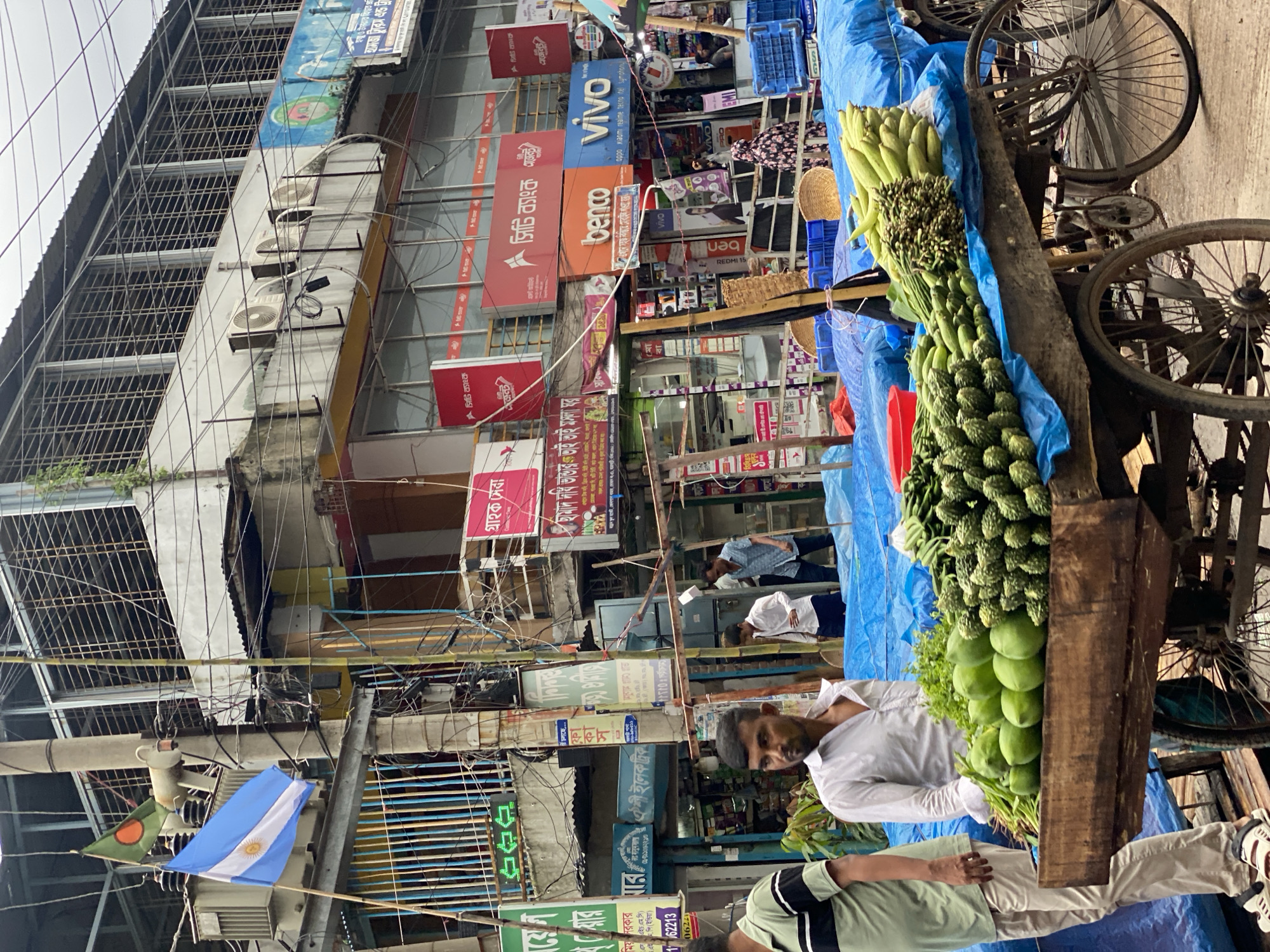 An outdoor scene in Dhaka, Bangladesh, features a street market. In the foreground, a man stands next to a cart laden with fresh produce, including bitter gourds, green mangoes, and corn. The cart is covered with a blue tarp. Behind the cart, a man in a white shirt looks to the side, and further back, two men are walking. The background shows a multi-story building with various shop signs, some in Bengali. Flags, including an Argentinian flag and a Bangladeshi flag, are visible hanging from poles. The sky is overcast.