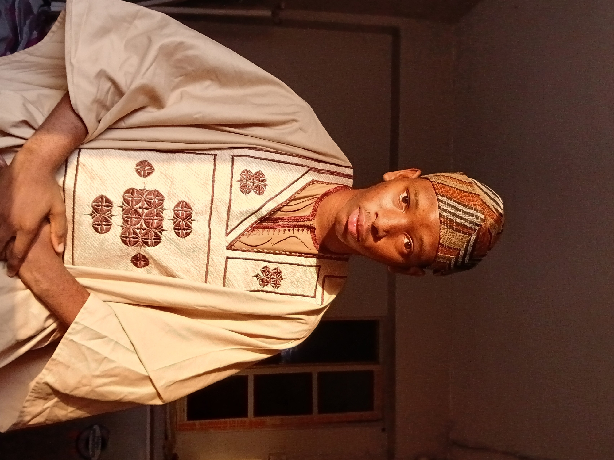 A medium shot depicts a man indoors. He wears a traditional attire consisting of a patterned brown and beige cap and a cream-colored robe with intricate embroidery. His hands are visible, positioned near his chest. The background shows a dark wall and a window with black squares. The location is not discernible from the image, so the claim that it is in Lagos, Nigeria, cannot be confirmed.