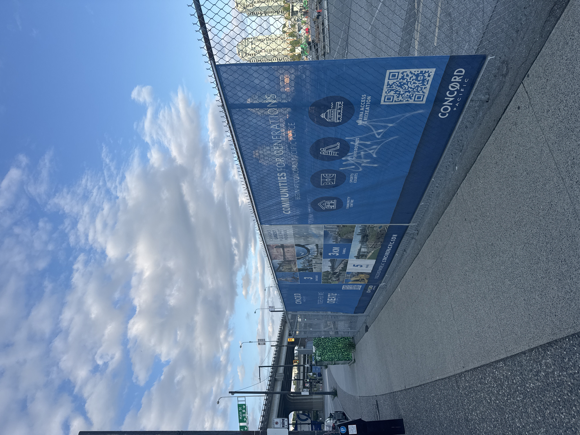The image depicts a construction site in Vancouver, Canada, specifically at Concord Pacific Place. A large blue banner on a chain-link fence advertises "COMMUNITIES FOR GENERATIONS" and highlights amenities like a community centre, sports courts, playgrounds, and marina access. The banner also features a QR code and the Concord Pacific logo. In the background, tall residential buildings and a highway overpass are visible. A sidewalk runs alongside the fence, with a street and a parked car to the left. The sky is partly cloudy.