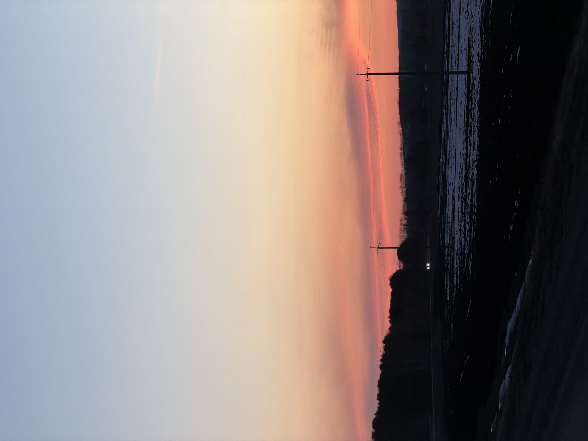 The image depicts a sunset over a rural landscape, possibly in the vicinity of Eldushino, Russia. The sky transitions from pale blue on the left to shades of orange and pink towards the horizon. Two electrical poles with wires are silhouetted against the vibrant sky. The horizon features a dark treeline. To the right of the treeline, a dark, reflective body of water or flooded field is visible. A road runs along the lower right corner of the image, also obscured in shadow. The overall scene is dim, characteristic of twilight or dusk. Two light sources (possibly car lights) are vaguely visible in the background near the treeline.