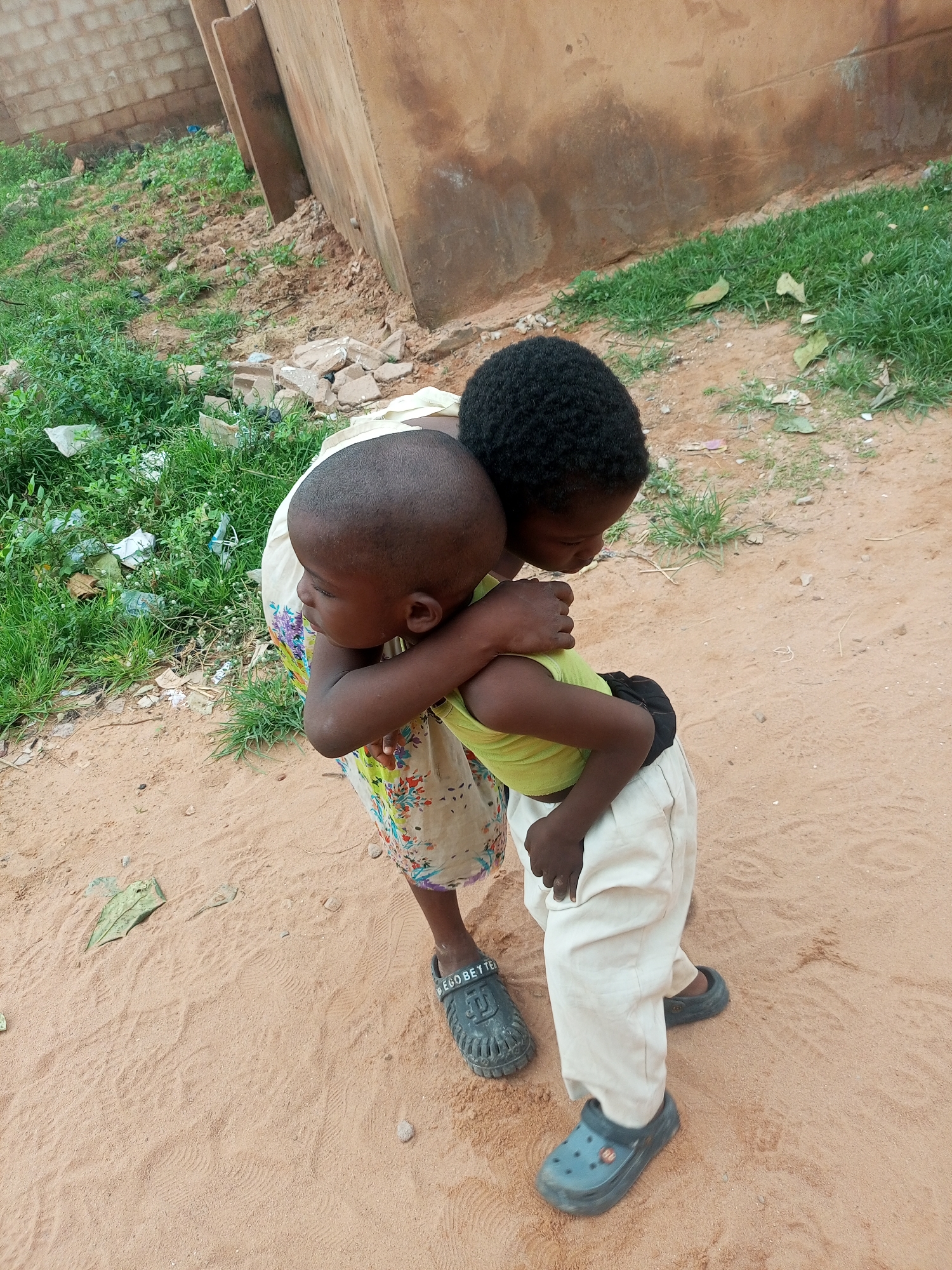 In Tempure, Nigeria, two young children are embracing outdoors on a sandy, dirt-covered ground. The child in the foreground, facing away from the camera and to the left, has a shaved head and is wearing a light-colored, floral-patterned skirt and a bright yellow t-shirt. They are being hugged from behind by another child, whose dark curly hair is visible. This second child is wearing light-colored pants and dark blue Crocs. The background shows a dusty, brown wall or building and patches of green grass with some scattered debris and plastic. The overall scene suggests a casual, outdoor environment, likely in a rural or less developed urban area.