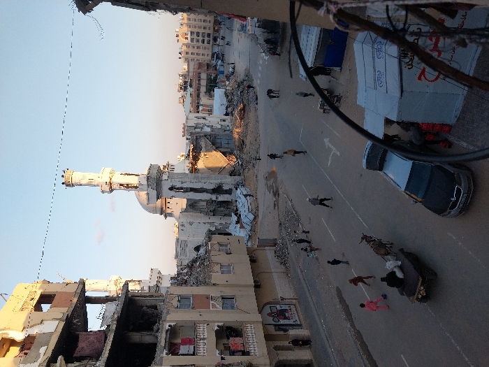 This image captures a street scene in Khan Yunis, Palestinian Territory, characterized by extensive urban damage under a clear sky.
In the midground, a large mosque with a central dome and two minarets is prominently featured; both minarets and the mosque's base display severe structural damage and fragmentation. To the immediate left of the mosque, a multi-story building exhibits significant damage, including exposed interiors, fractured facades, and visible impact marks. Laundry is observed hanging on balconies of this building. Debris is scattered across the ground surrounding the mosque and extending into the paved street.
The street, marked with white lines and a directional arrow, shows signs of disarray. Numerous individuals, including adults and children, are present on the street and sidewalks, walking or standing. In the lower left, a donkey-drawn cart carries at least one person and white sacks, moving along the street. On the right side of the street, a silver Skoda sedan is parked with its trunk open. Adjacent to the car is a white tent structure with red Arabic script and blue fabric details, with several individuals gathered nearby. Other damaged or partially constructed buildings are visible in the background.